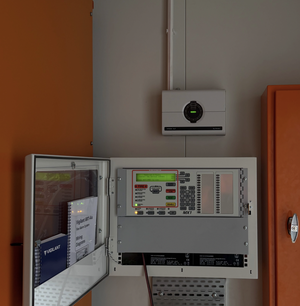 Fire alarm control panel and fire suppression system in a room with orange and white walls.