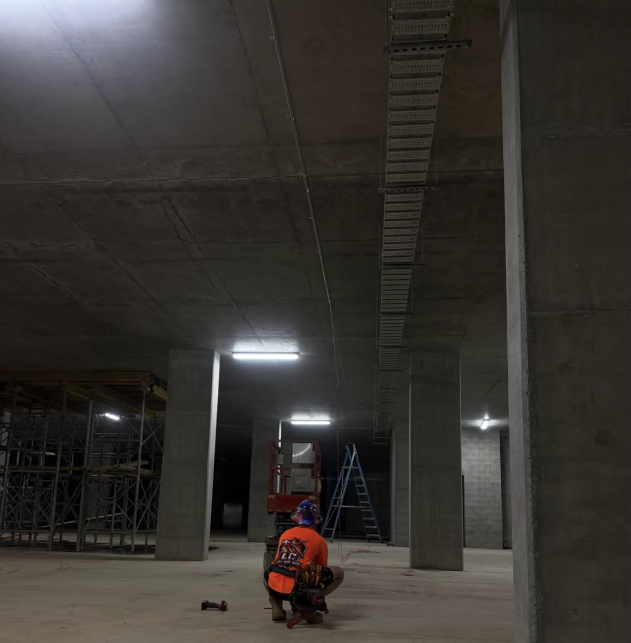 Construction worker kneeling on the floor in an unfinished building, with scaffolding, ladders, and construction equipment visible, and electrical conduit and cable trays installed on the ceiling.