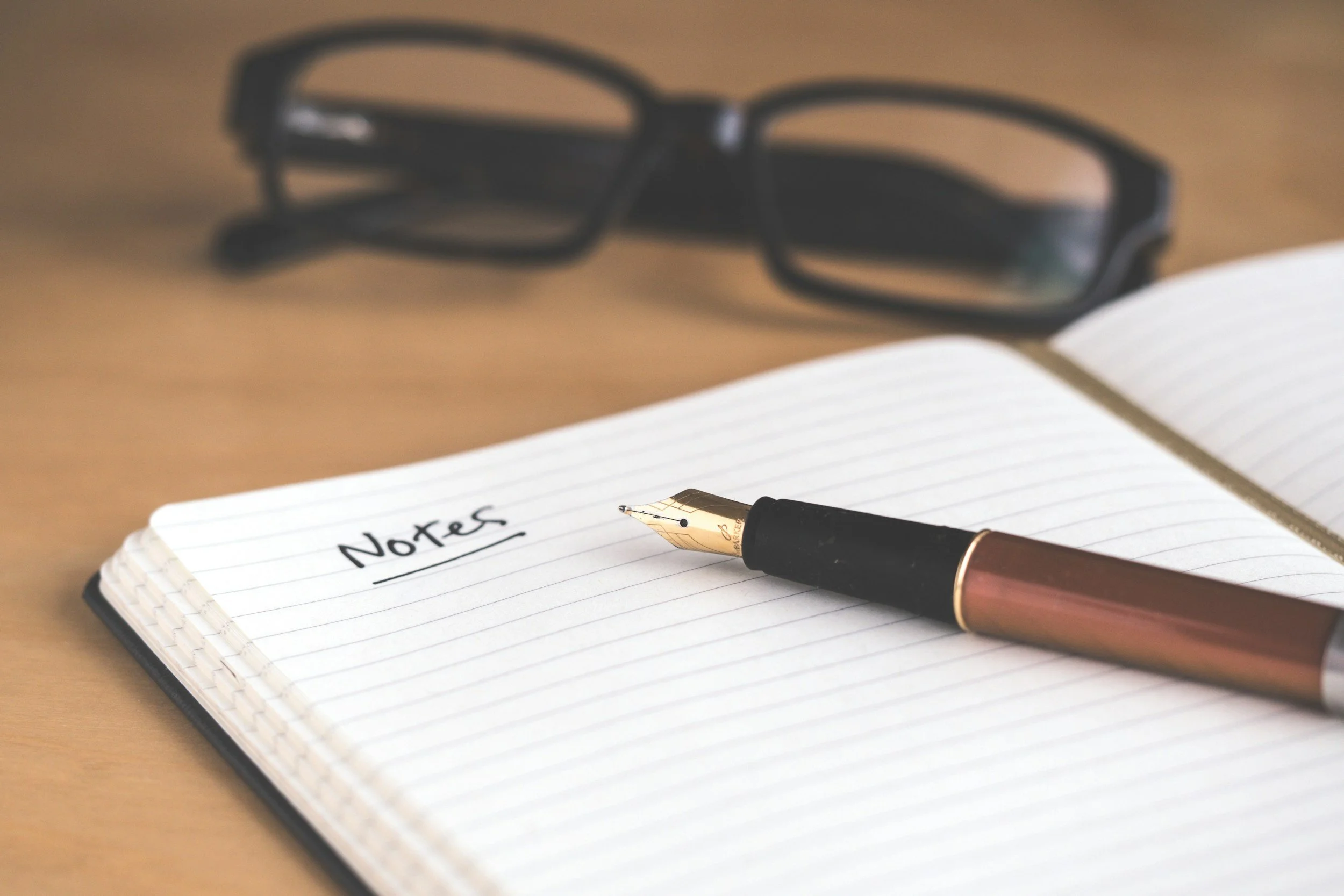 An open notebook with handwritten text that says "Notes," a fountain pen resting on the paper, and a pair of black glasses in the background, all placed on a wooden surface.