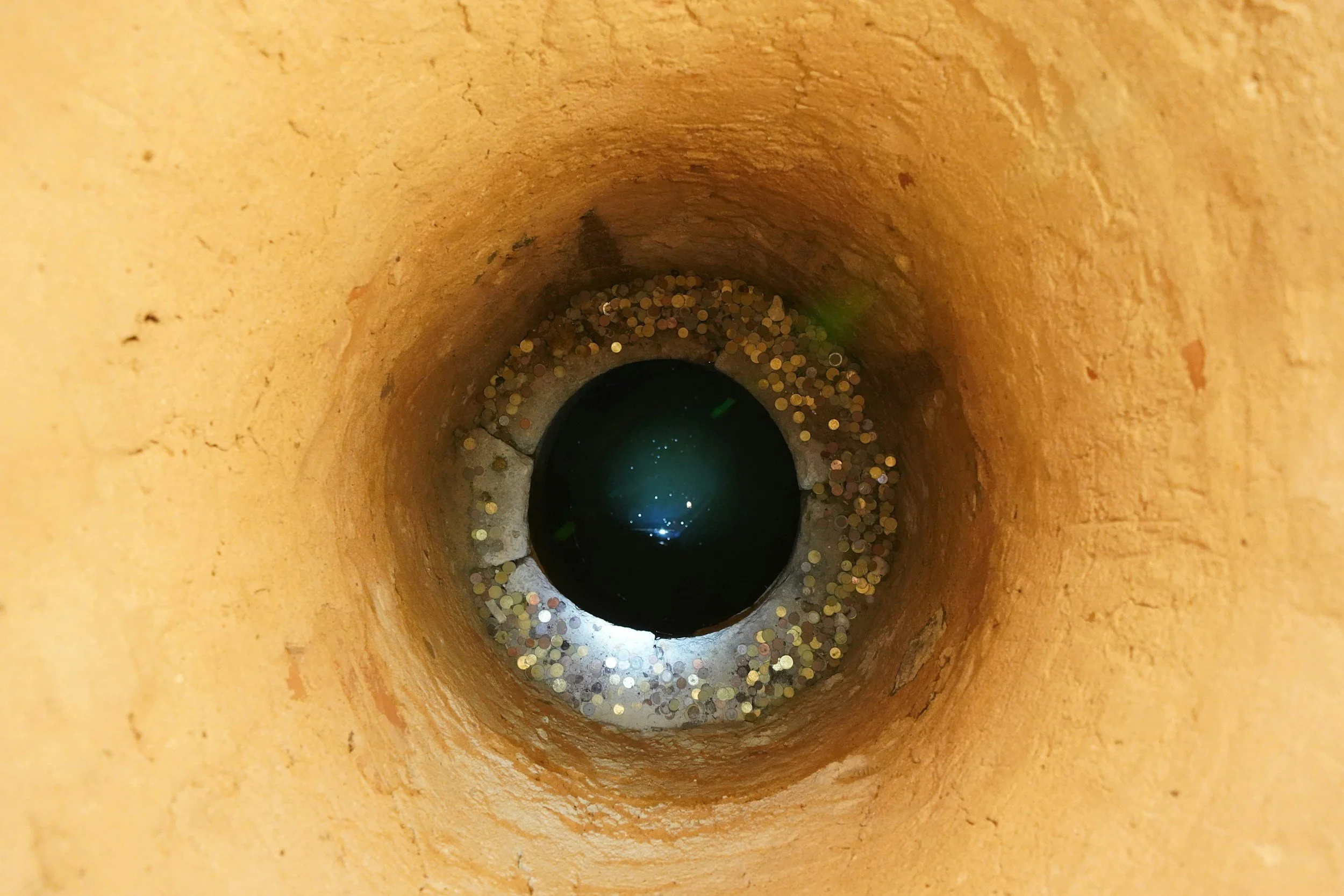 Inside of a yellow cylindrical pipe with coins and some debris at the bottom, and water reflecting light at the end of the pipe.