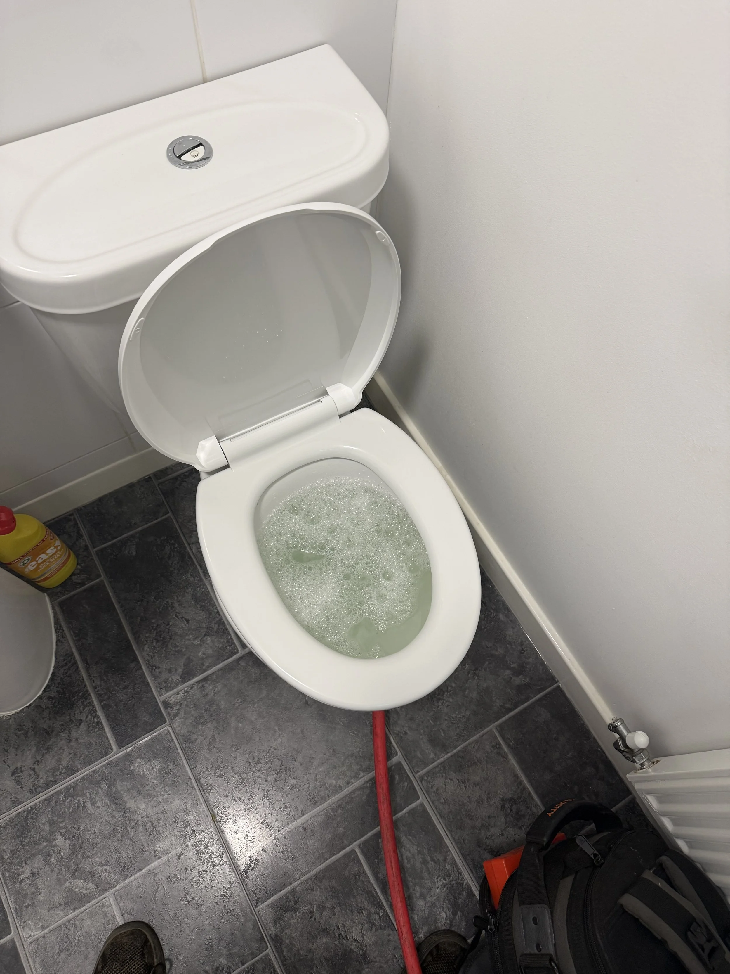 A white toilet with the lid up, filled with water. The toilet is in a bathroom with dark gray tiled floor and a white wall. Part of a yellow cleaning spray bottle and a black bag are visible in the foreground.