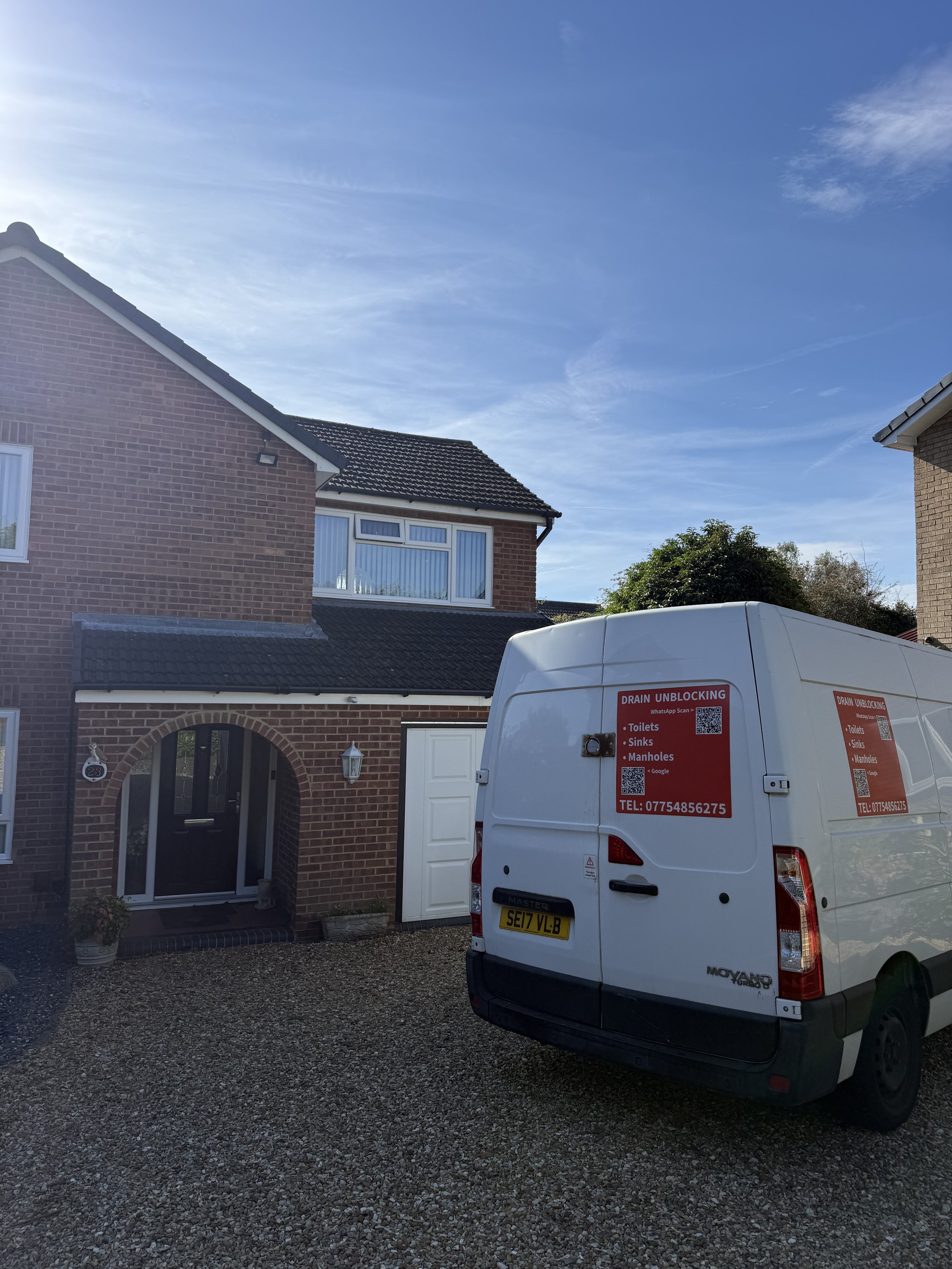 A white utility van parked in front of a brick house with a tiled roof, a white door, and a window with curtains. The house has a small front yard with gravel ground, and there is a bush on the right side. The vehicle has red and white signs indicating drain unblocking services.
