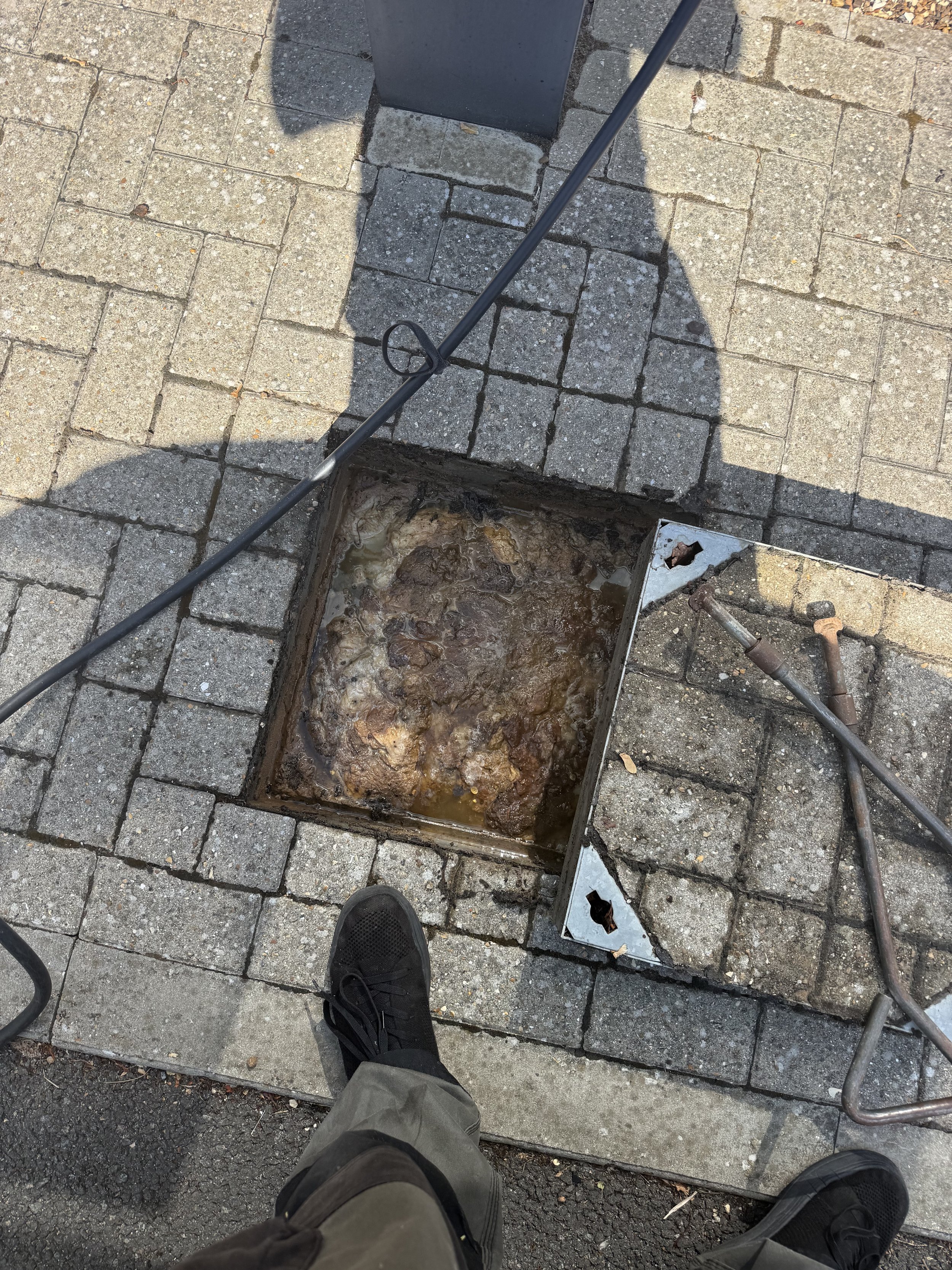 A sidewalk with gray bricks and a metal grating or cover open revealing rusted and damaged pipes and debris inside.