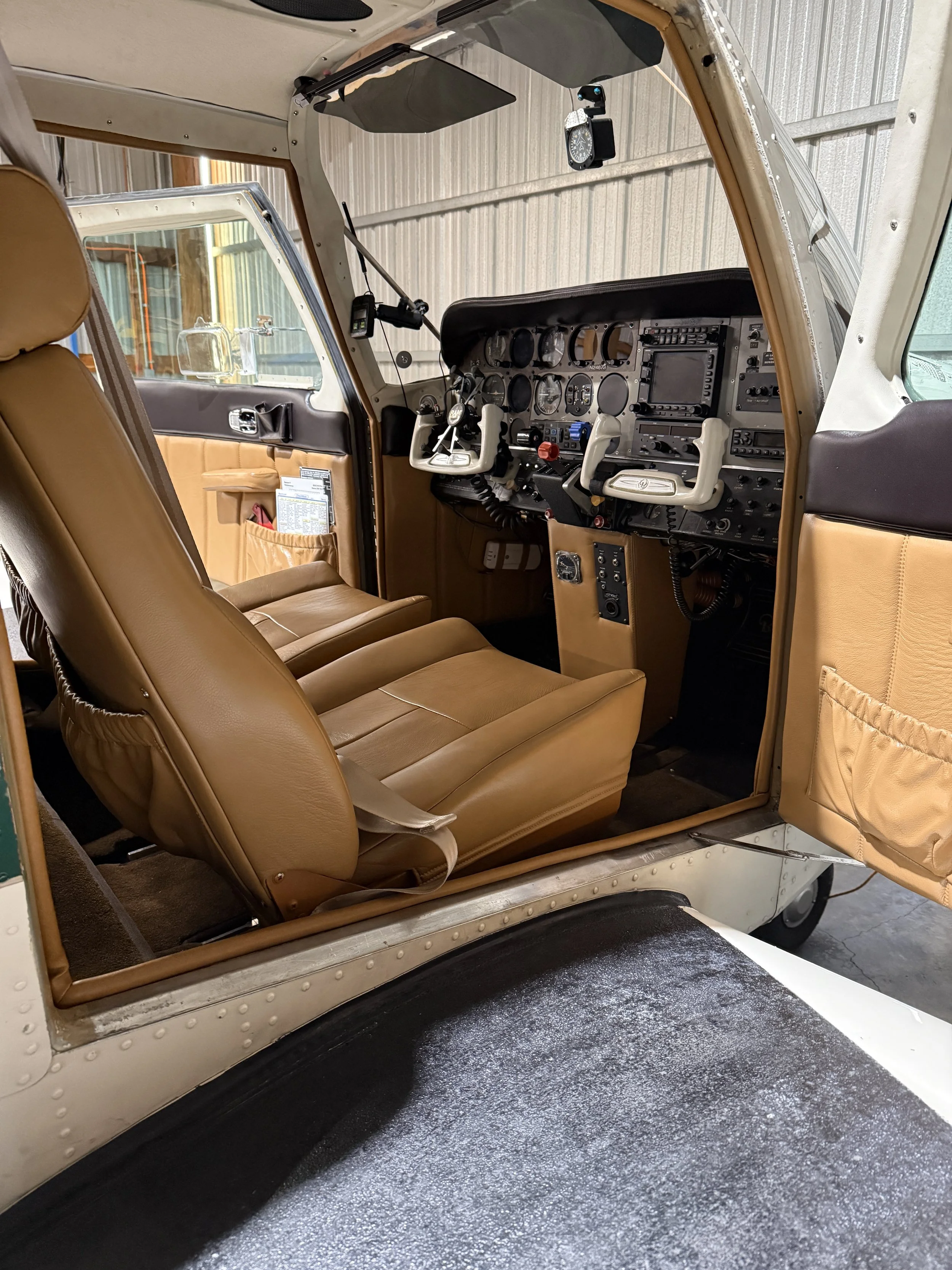 The interior of a small aircraft cockpit with tan leather seats, various gauges, controls, and radio equipment, and a clear view of the nose wheel through the open door.
