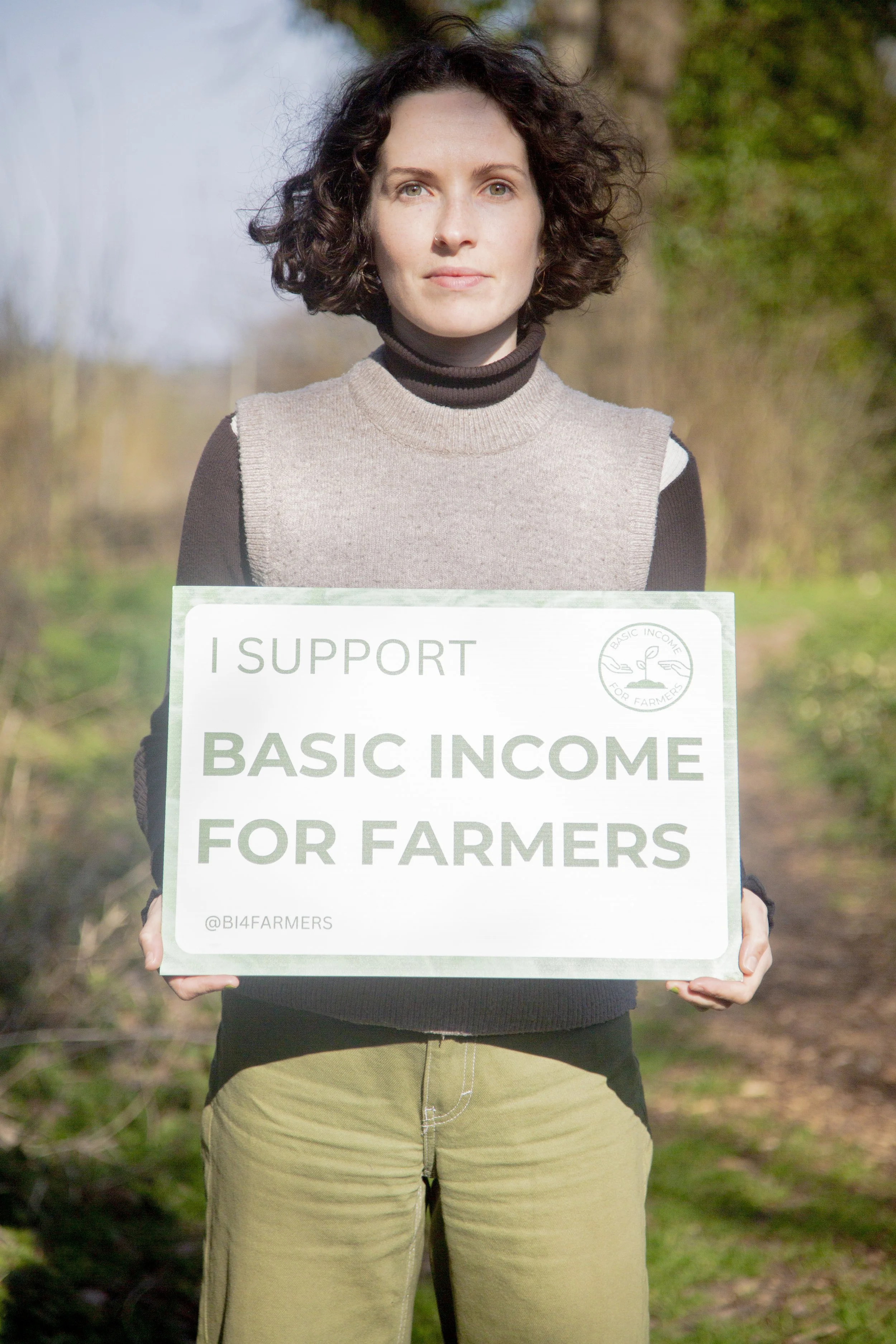 Jo Poulton, founder of Basic Income 4 Farmers. She is a white woman with short dark curly hair, standing outdoors holding a sign reading I Support Basic Income for Farmers.