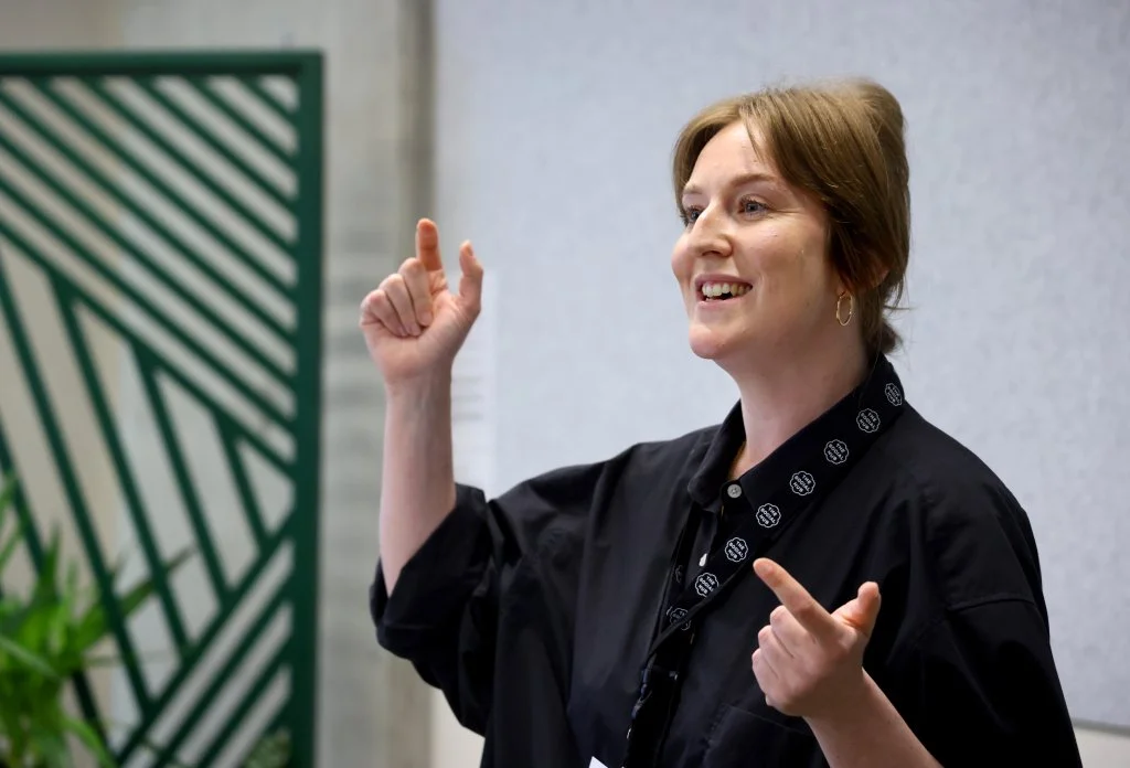 Cleo Goodman facilitating a session at The Melting Pot's Bridging Communities event in Glasgow. She has auburn hair tied back and is wearing a black shirt, gesturing expressively while speaking.