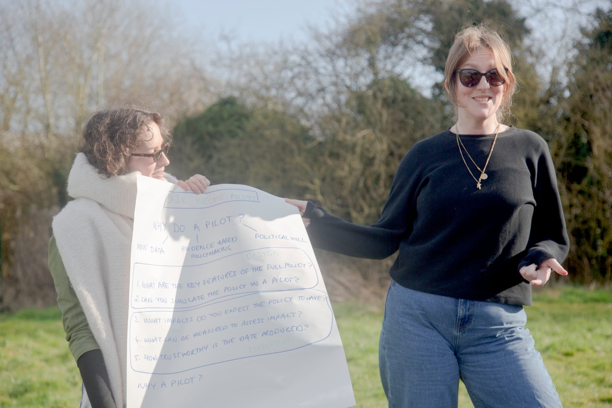 Cleo Goodman and Jo Poulton running a workshop on designing a basic income pilot. They are outdoors in the sun holding a big piece of flipchart paper.