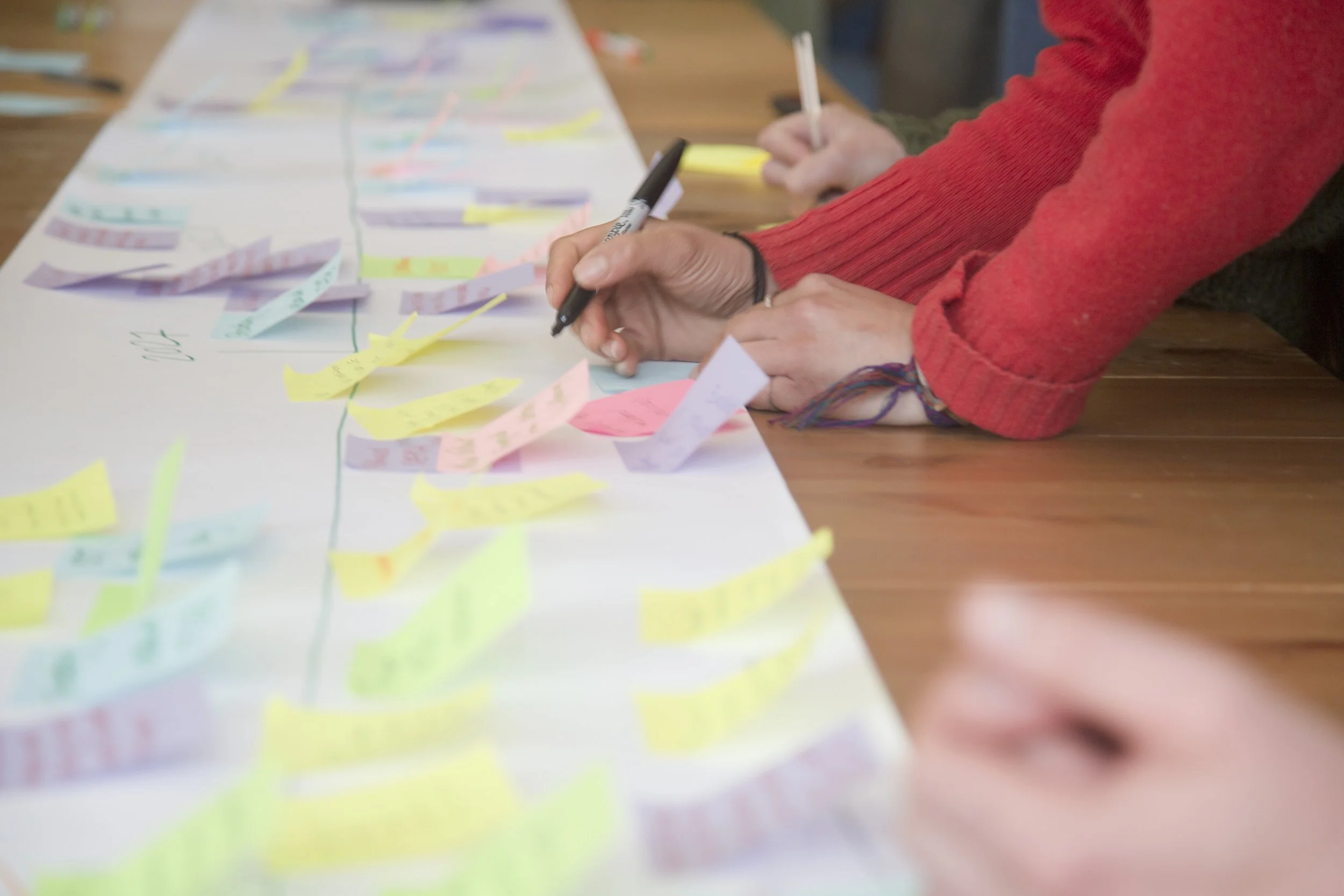 Close-up of participants' hands writing on colourful sticky notes arranged on a long paper timeline during a participatory workshop.
