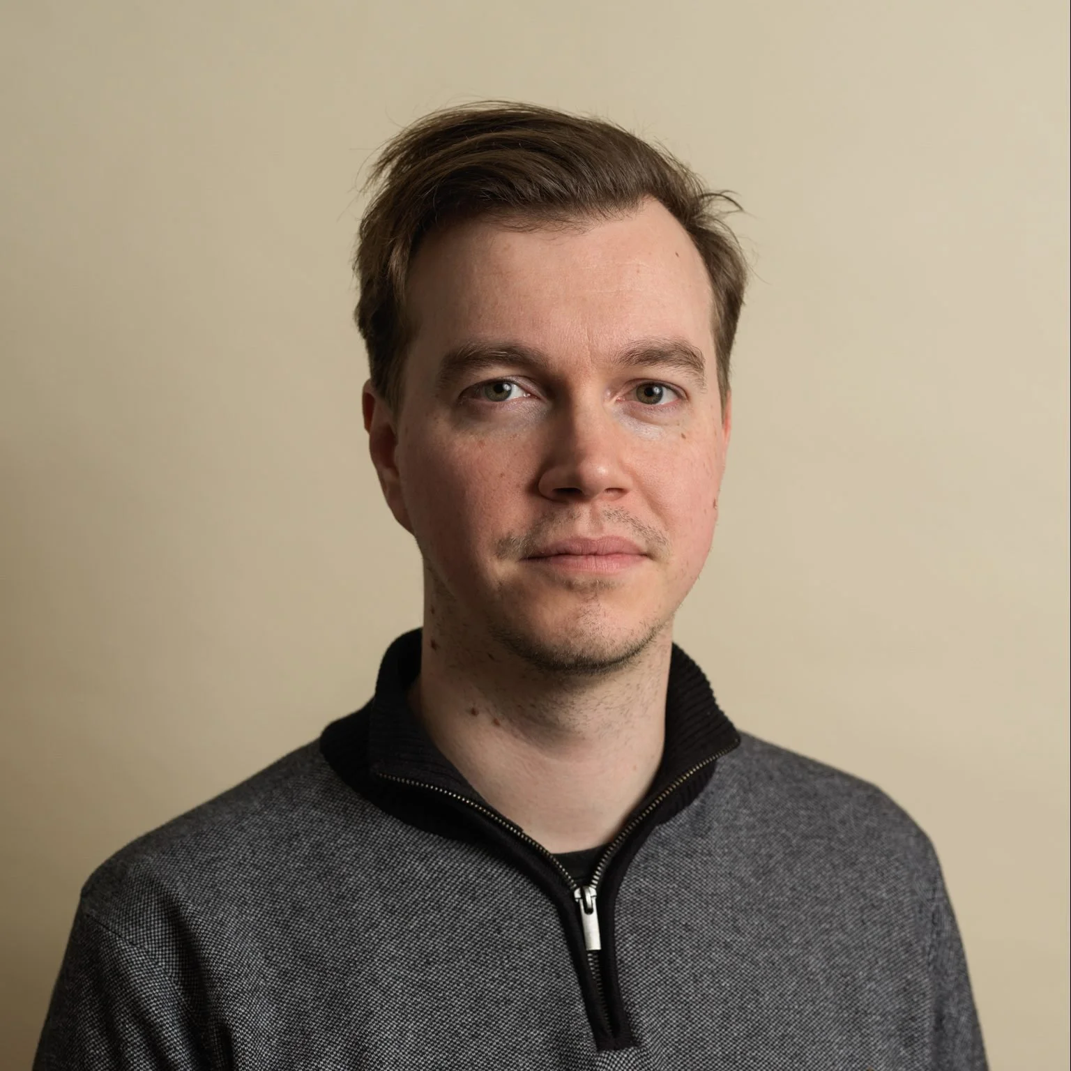 A portrait of a young man with light skin, short brown hair, wearing a grey zip-up pullover with a black collar against a plain beige background.
