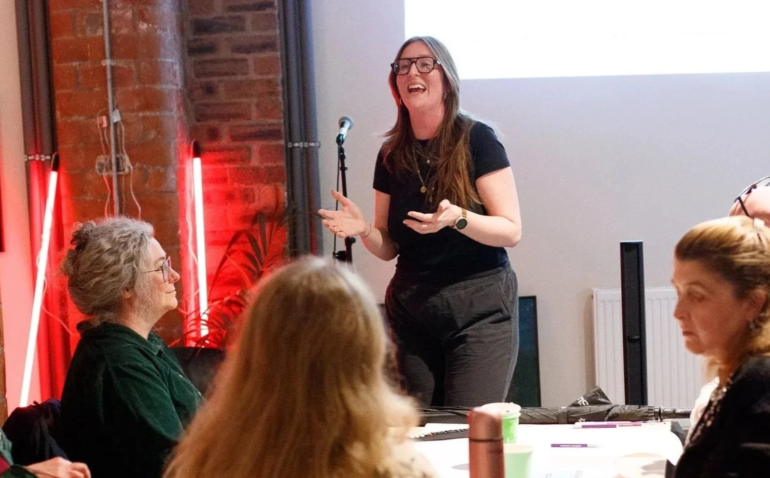 Cleo Goodman facilitating a workshop in a brick-walled venue with red neon lighting, gesturing expressively while speaking to a seated group of participants. She is a white woman with long auburn hair wearing glasses and a black top.