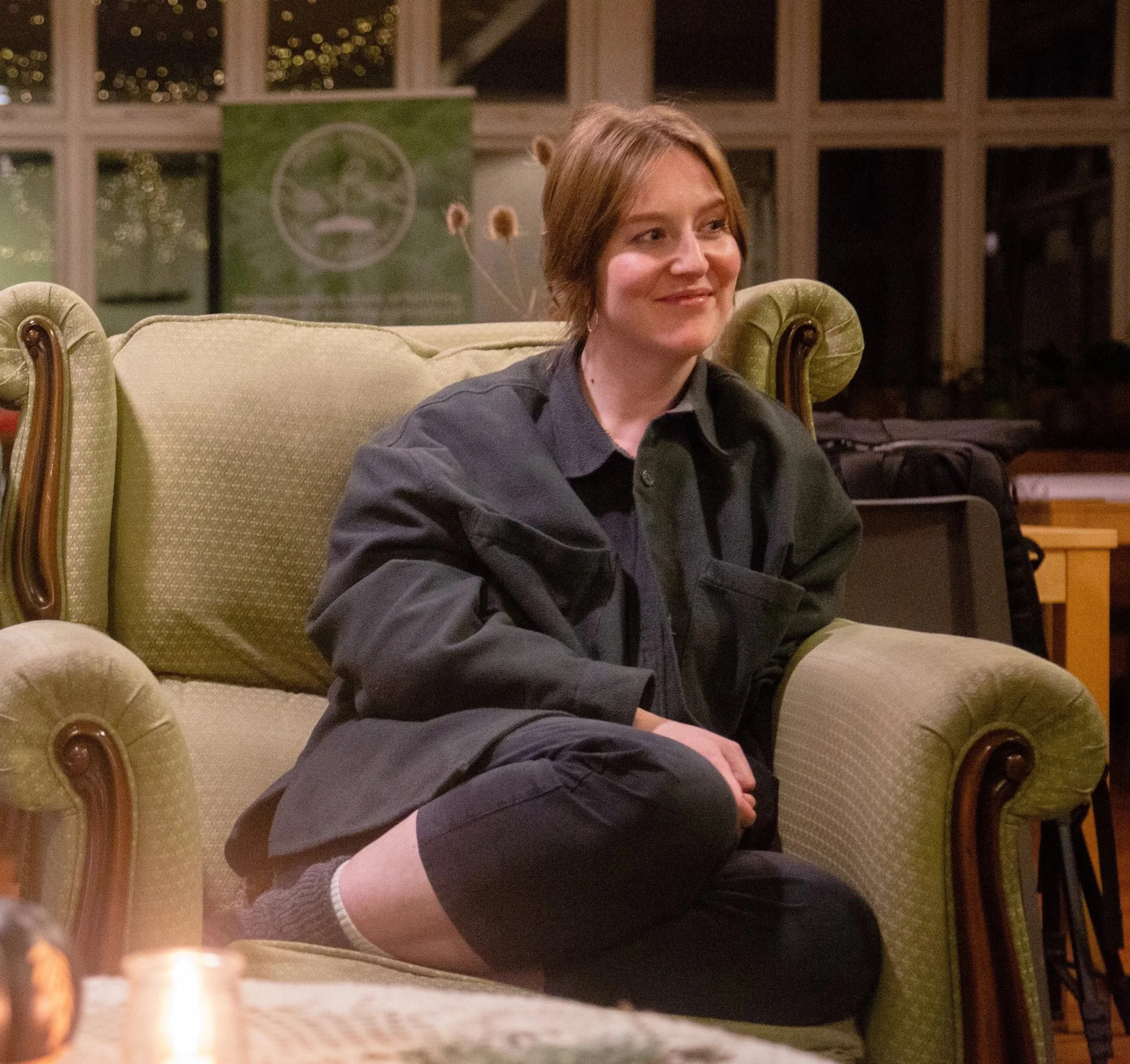 Cleo Goodman at the Basic Income 4 Farmers campaign strategy weekend. She is a white woman with fair auburn hair, wearing a dark jacket, sitting relaxed on a green sofa and smiling, with a campaign banner visible in the background.