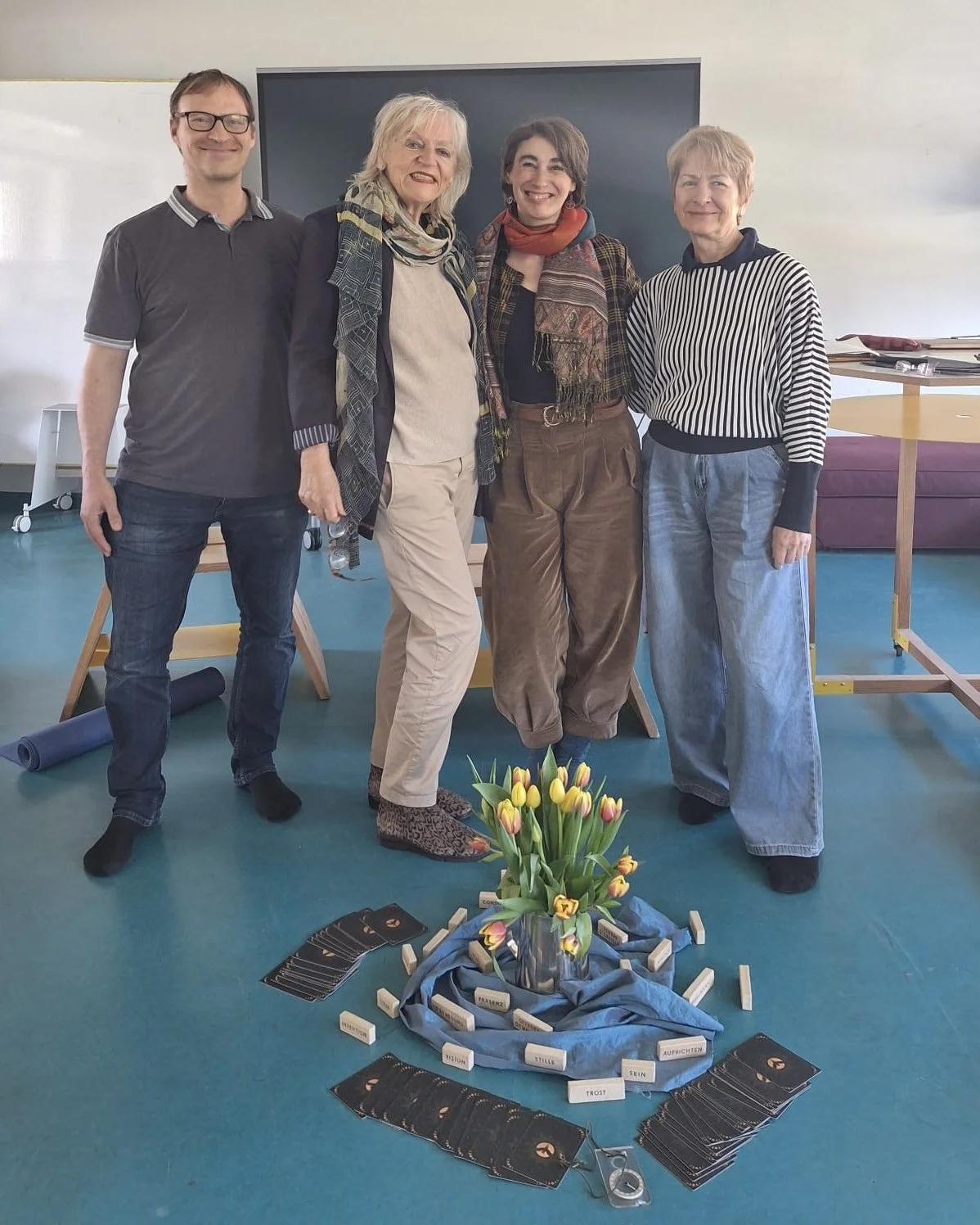 Vier Frauen und ein Mann stehen im Raum, vor einem schwarzen Display, mit einer Blumenvasen-Installation mit Tulpen, umgeben von Steinen, Karten und Gegenständen, vermutlich bei einer Gruppenaktivität oder einem Workshop.