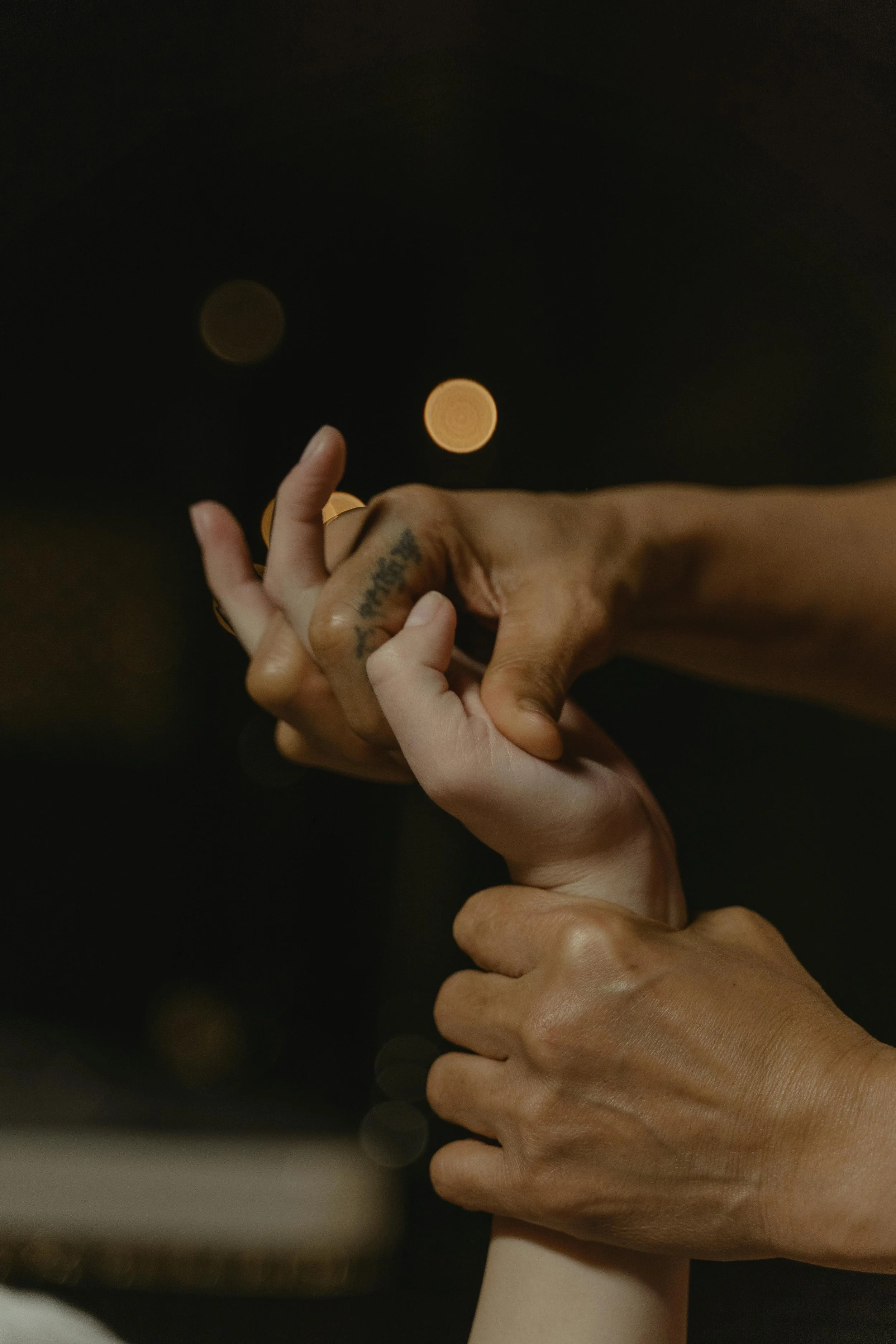 Two hands, one with a tattoo, exchanging a small object against a dark background with blurred orange lights.