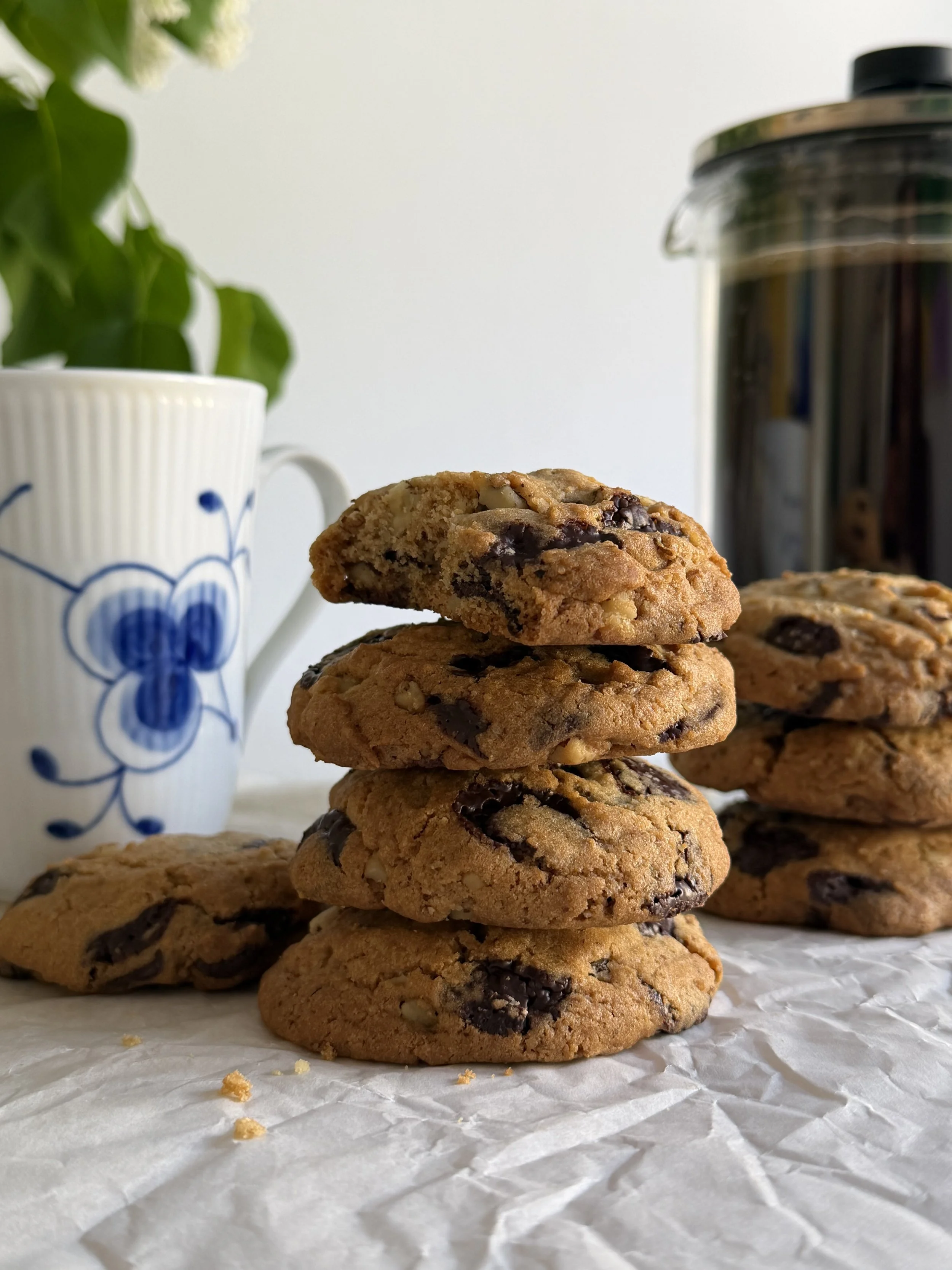 Stack of chocolate chip cookies on crinkled white paper, with a cup with blue floral design and a plant in a white pot in the background.