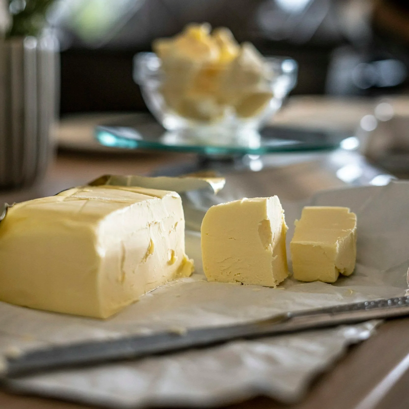 Uncut block of butter with several slices removed, placed on a tray. Out of focus in the background is a glass dish with scoops of butter.