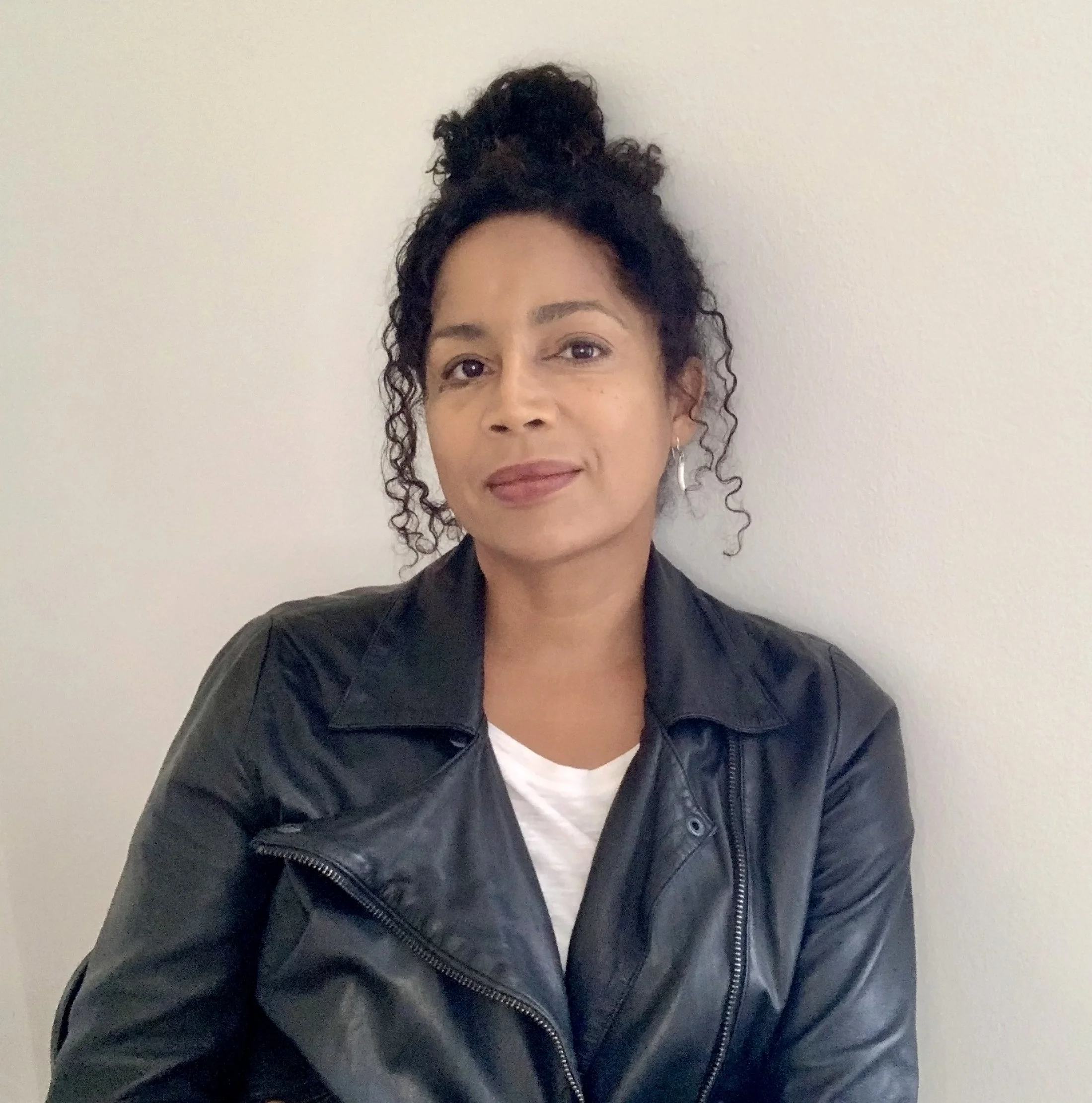 A woman (founder Holly McWhorter) with curly dark hair tied up in a bun, wearing a black leather jacket and a white shirt, standing against a plain white wall.