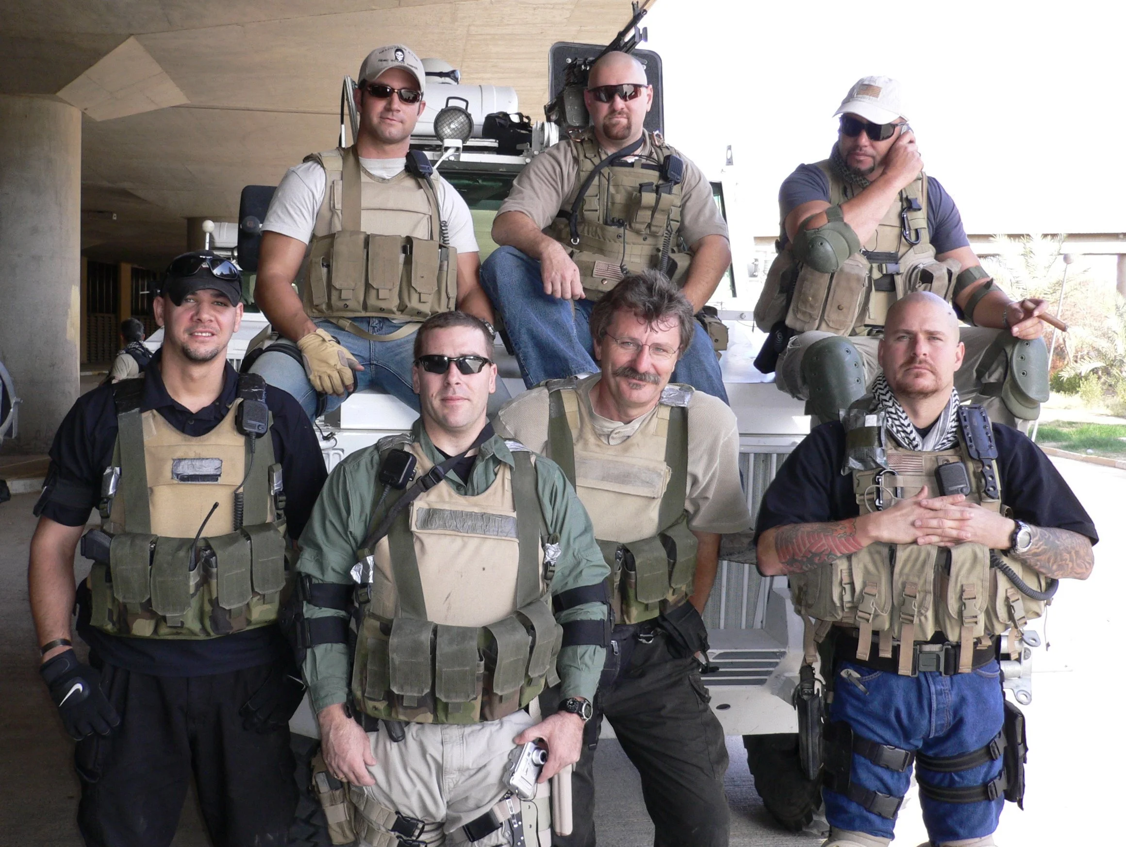 Group of seven men posing outdoors, wearing tactical gear and sunglasses, with some holding guns, in front of a building or structure.