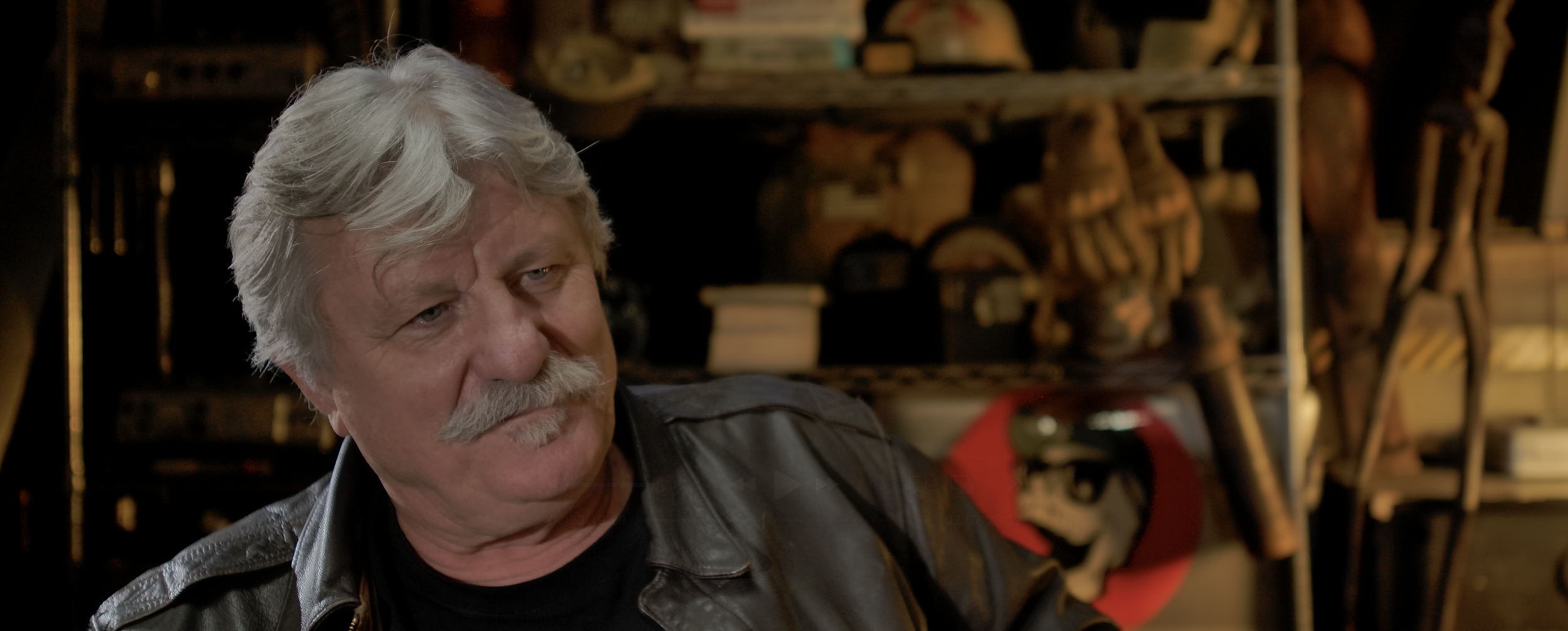 Older man with gray hair and mustache wearing black leather jacket, sitting indoors with shelves of shoes and books in the background.