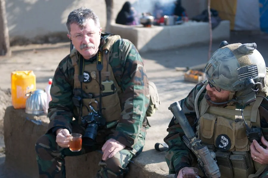 Two military personnel in camouflage uniforms sitting outdoors, one holding a glass of tea or coffee, with other gear and supplies around them.