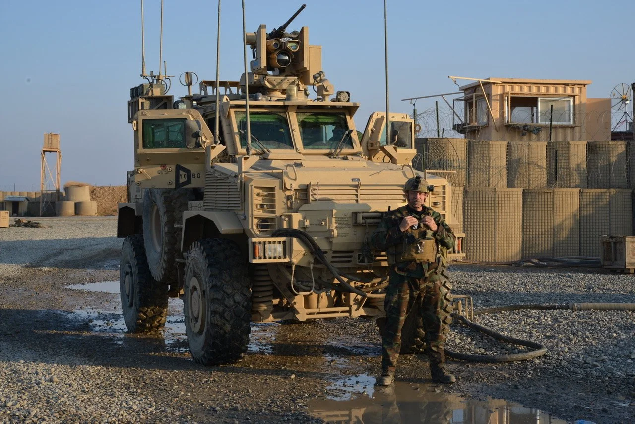 A soldier in camouflage uniform standing next to a tan military vehicle in a desert military base, with a watchtower and sandy structures in the background.