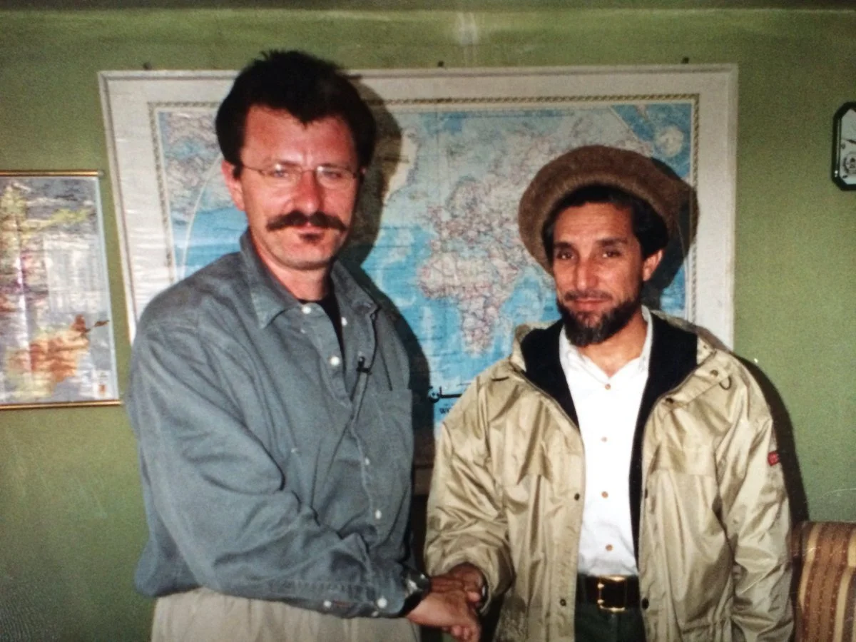 Two men shaking hands in front of a world map on the wall, with additional framed pictures on a green wall.