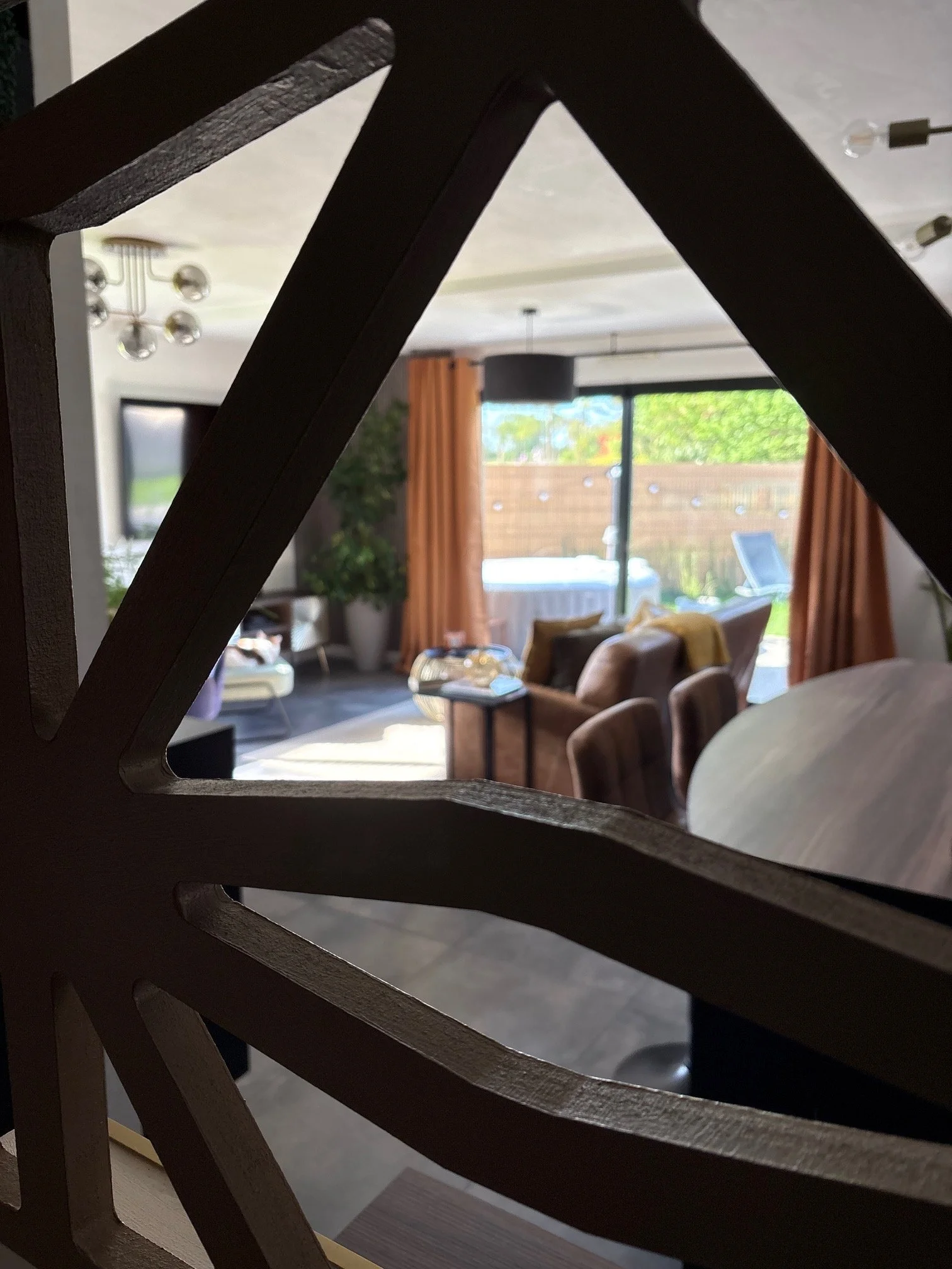 Vue à travers une structure en bois d'une pièce de salon avec des meubles, une grande fenêtre, des rideaux oranges, et des éléments décoratifs.