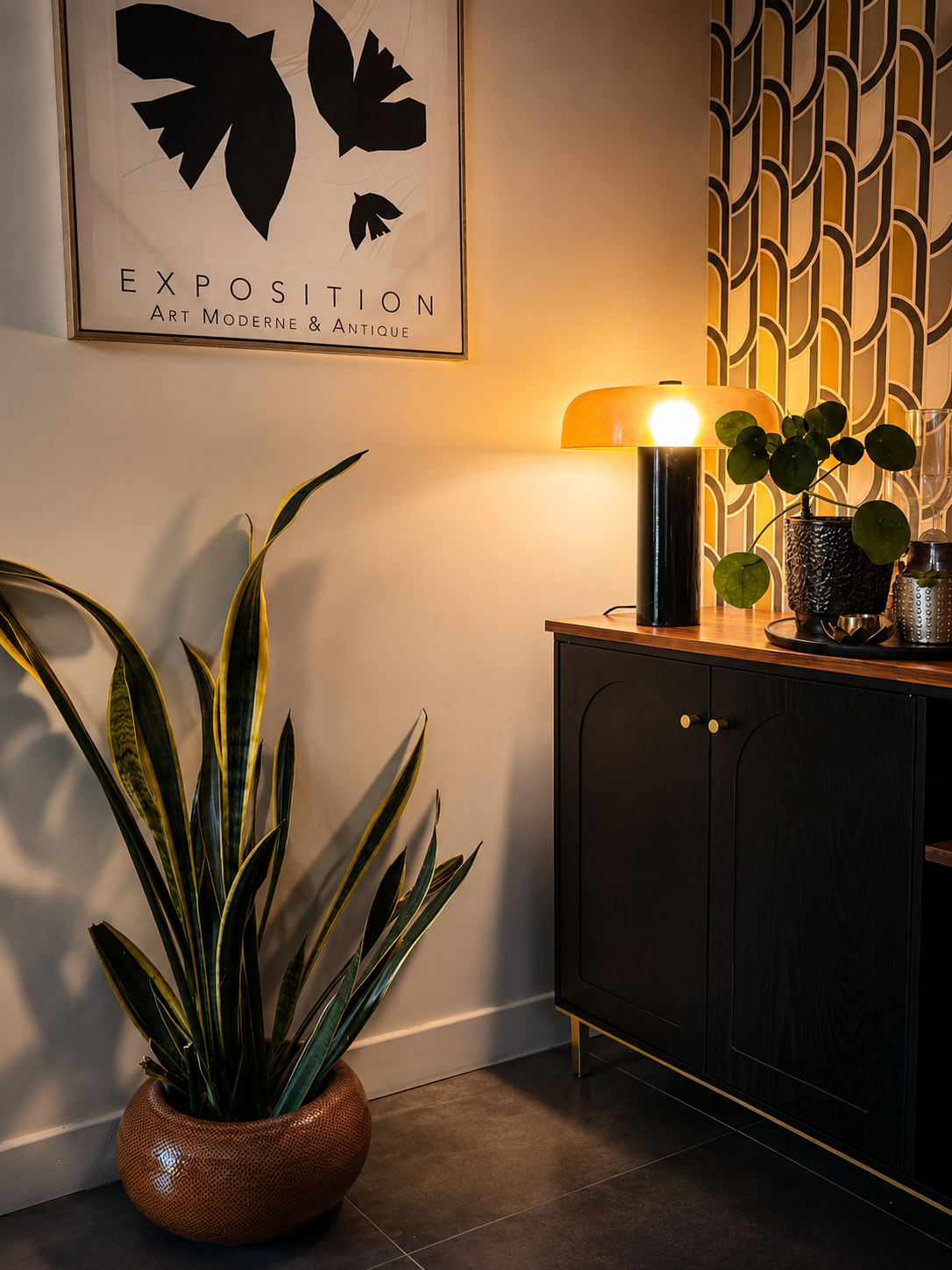 Intérieur moderne avec une plante verte dans un pot en rotin, une commode noire avec une surface en bois, et une lampe sur la meuble. Un tableau avec le texte 'Exposition Art Moderne & Antique' est accroché au mur, avec un papier peint à motifs géométriques à l'arrière.