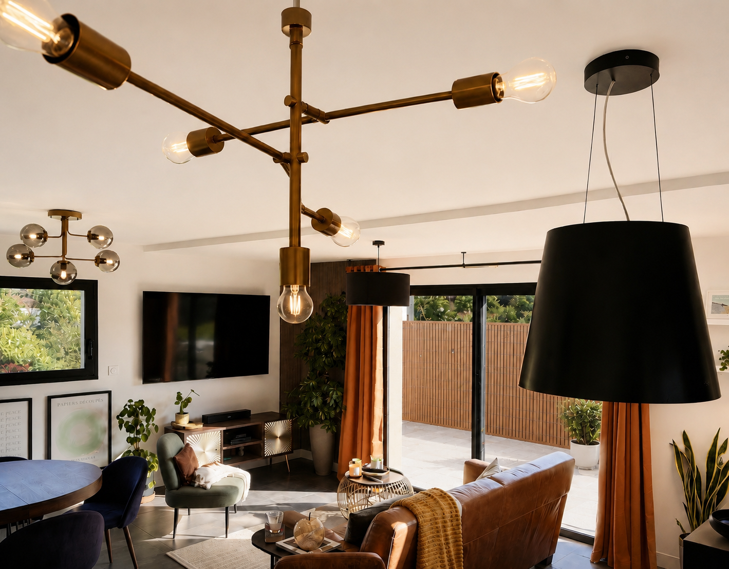 Intérieur d’un salon moderne avec luminaires suspendus, un téléviseur mural, des plantes vertes, un canapé en cuir, une table basse en rotin, et de grandes fenêtres avec des rideaux orange.