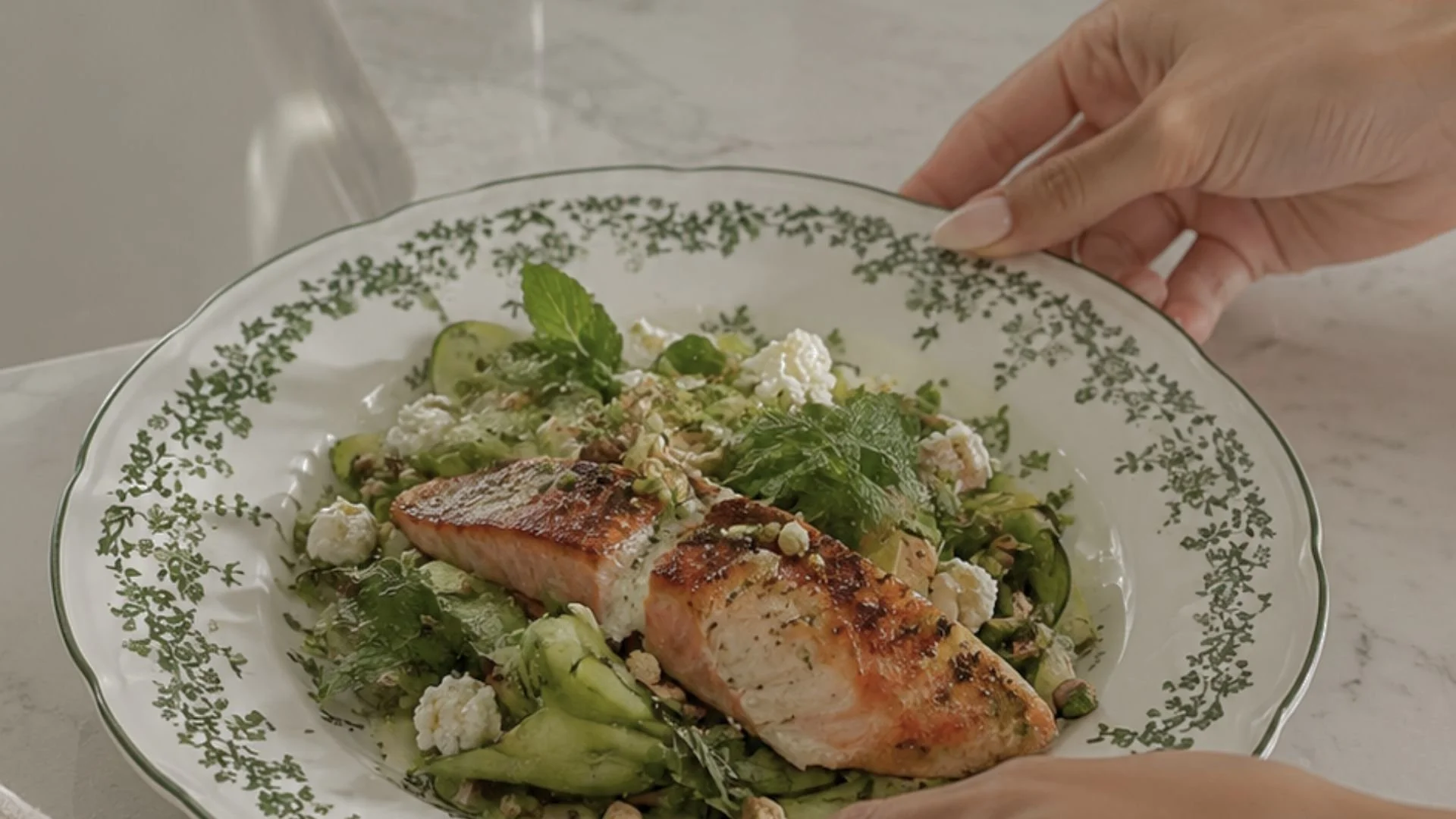 A plate of cooked salmon on a bed of salad with cucumbers, herbs, and cheese, garnished with mint leaves, on a decorative glass plate being held by a hand.