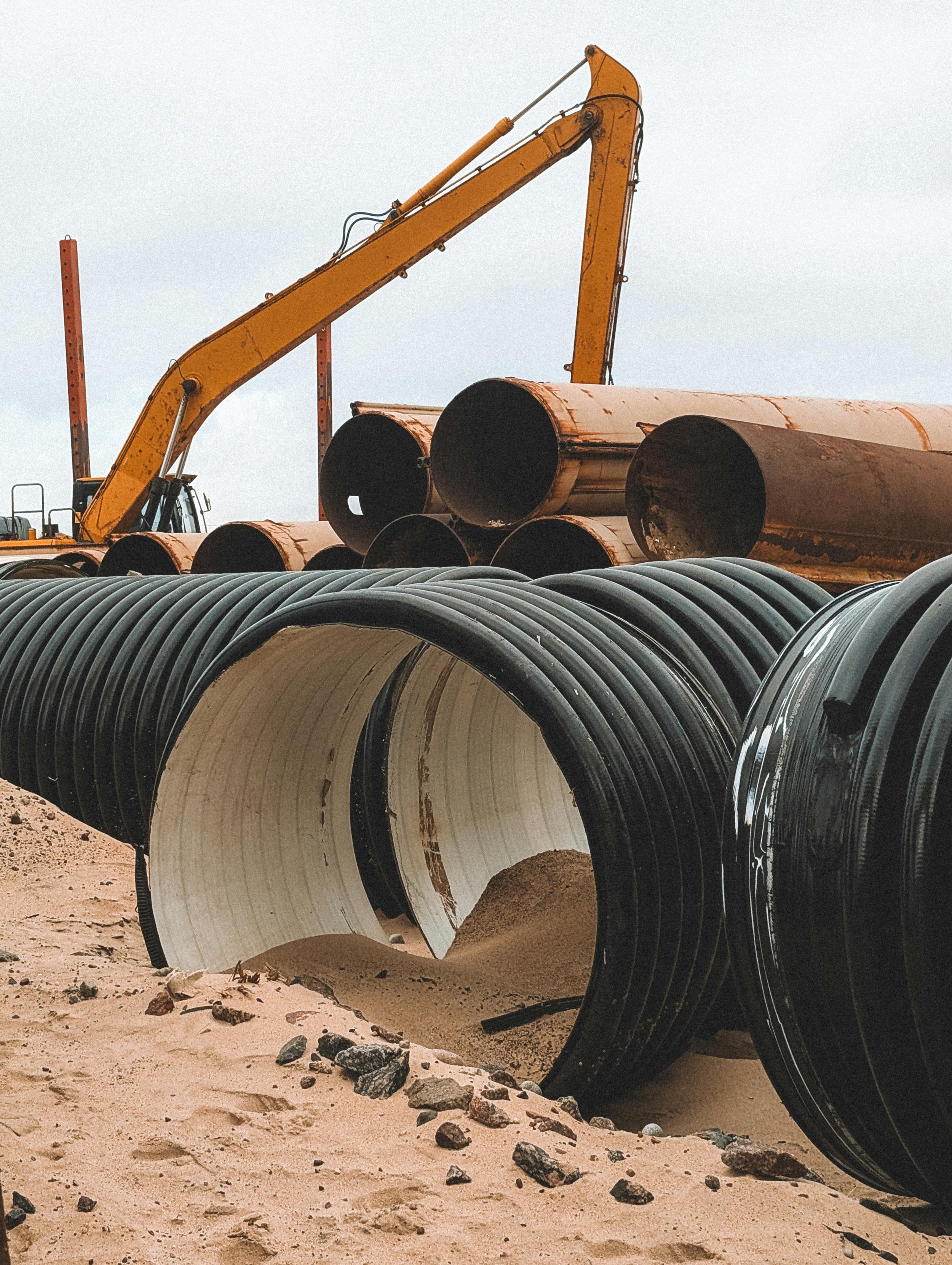 Construction site with large pipes laid on sandy ground, and a yellow excavator in the background.