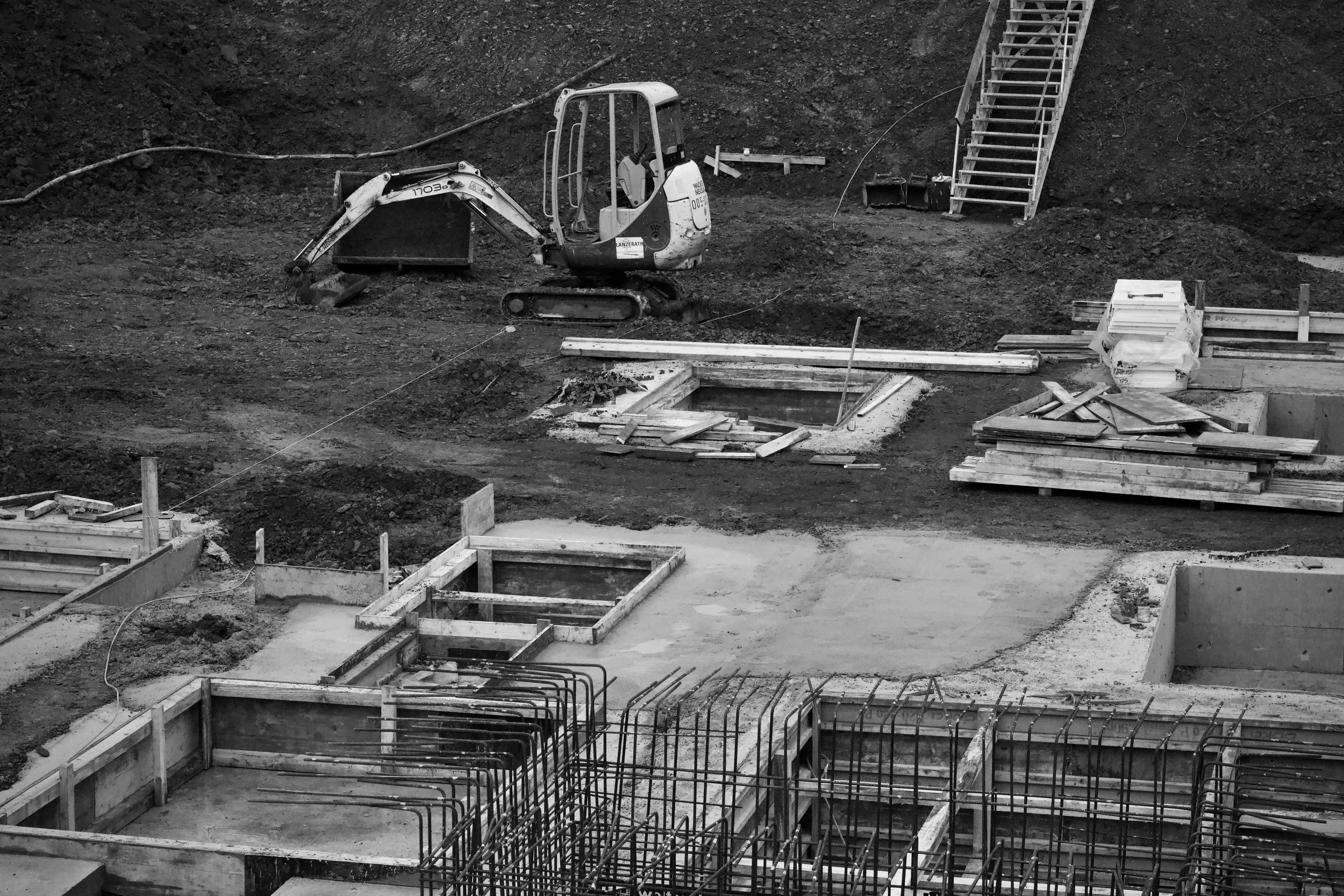 Black and white photo of a construction site with a small excavator, wooden forms, and rebar for concrete