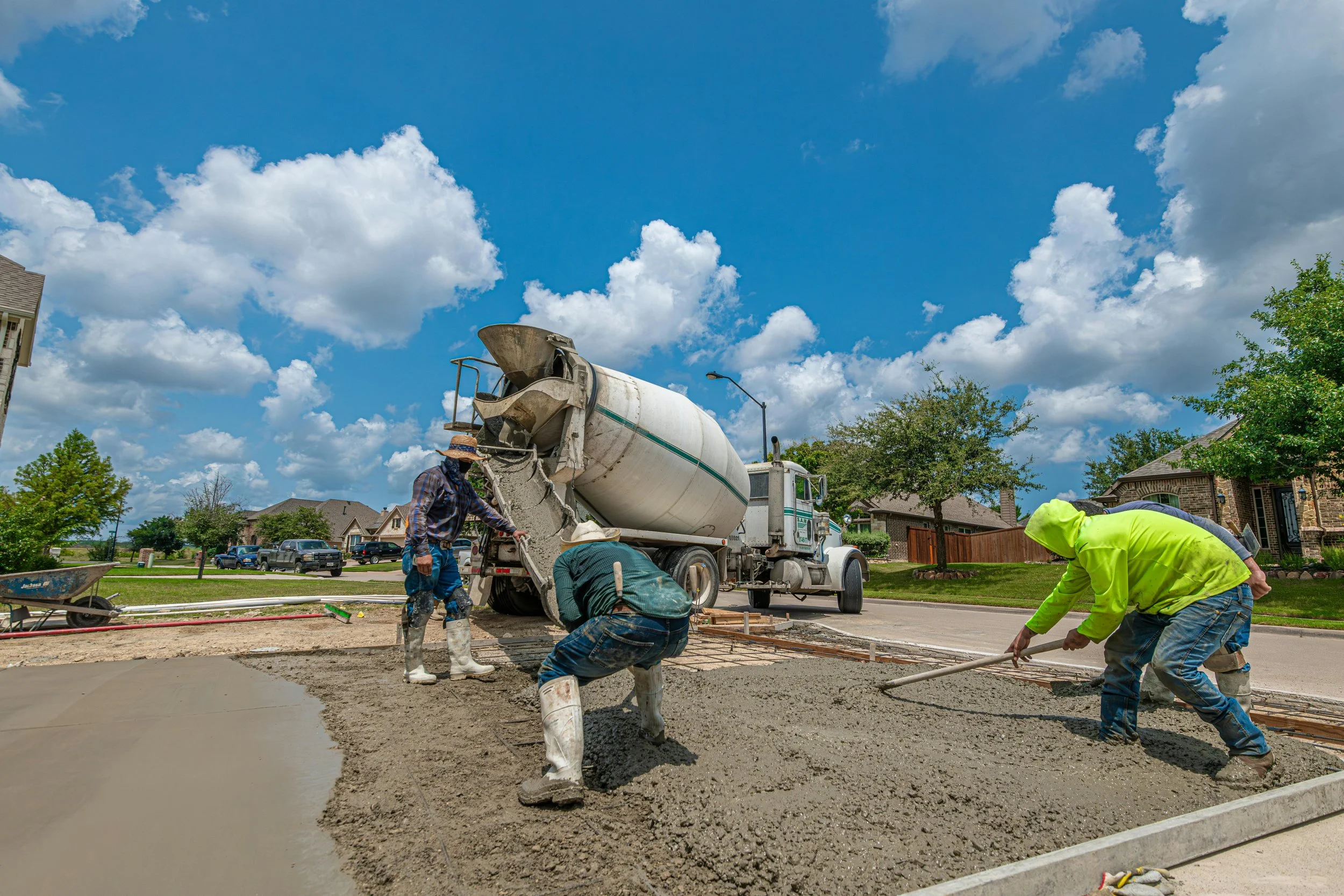 Workers are pouring and leveling concrete for a sidewalk or driveway in a residential neighborhood on a sunny day.