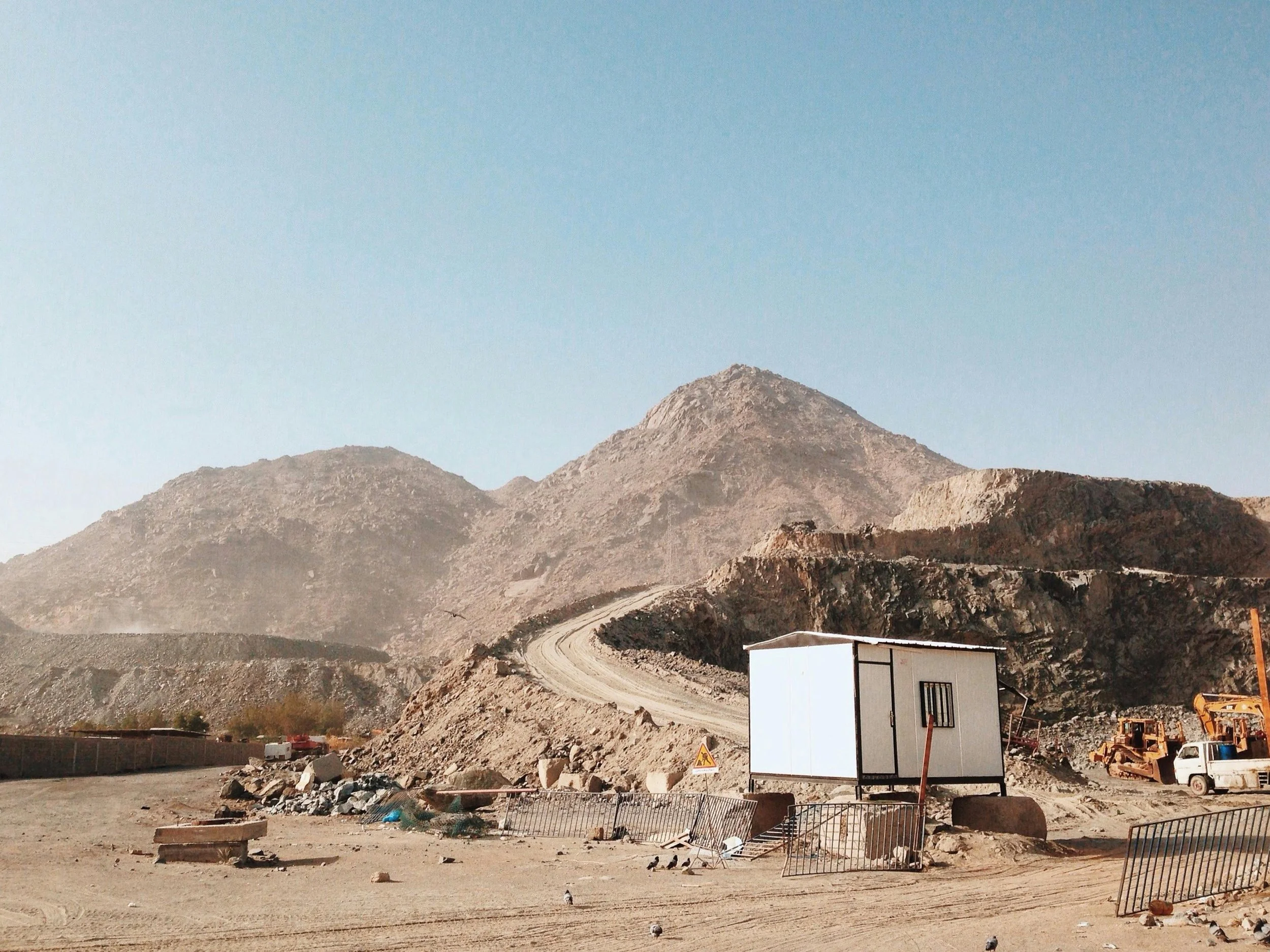 A construction site at the base of arid, rocky mountains under a clear sky, with a small white building, construction equipment, and a dirt road.