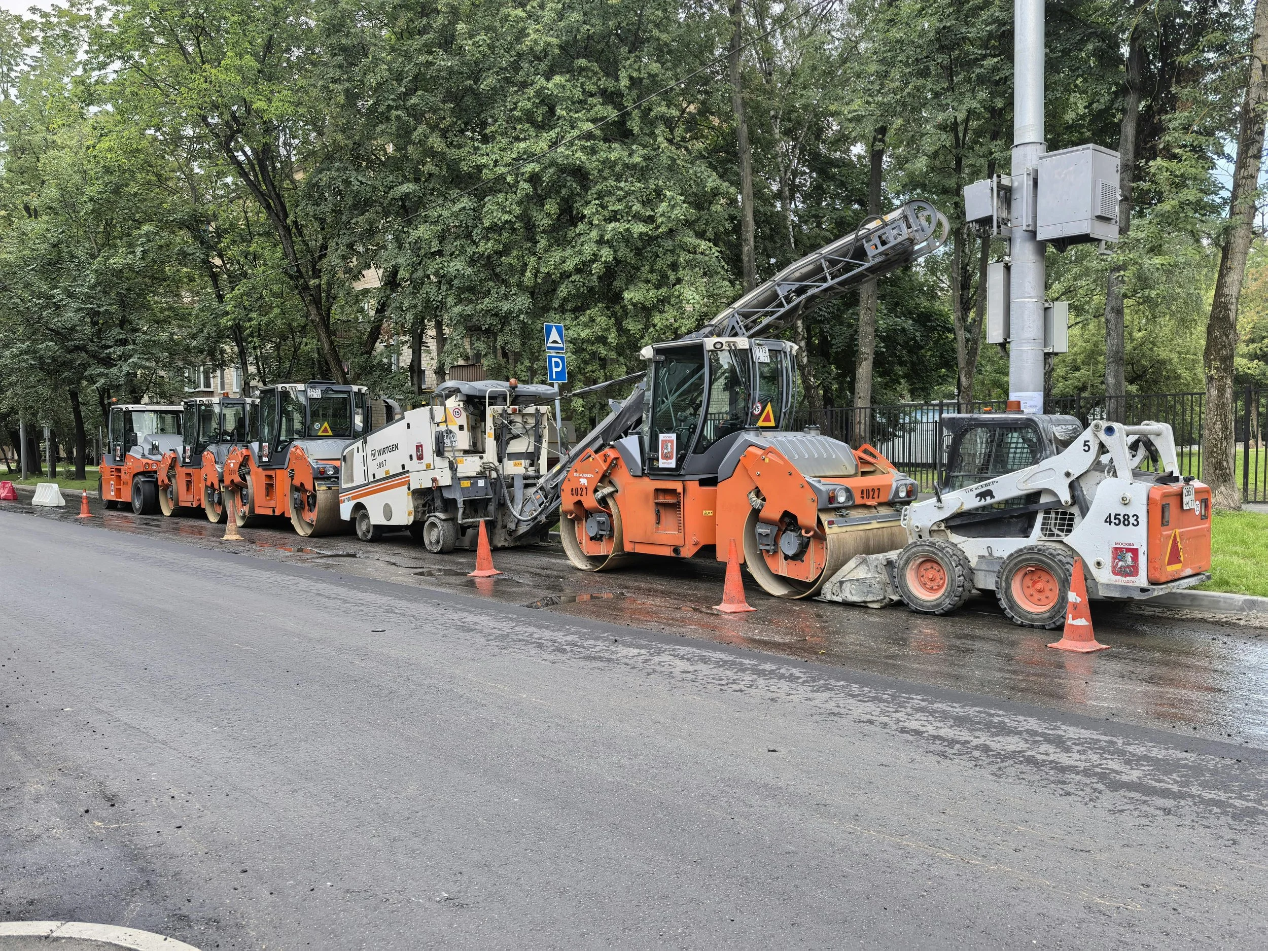 Road construction with paving machines and orange cones near a park with green trees.