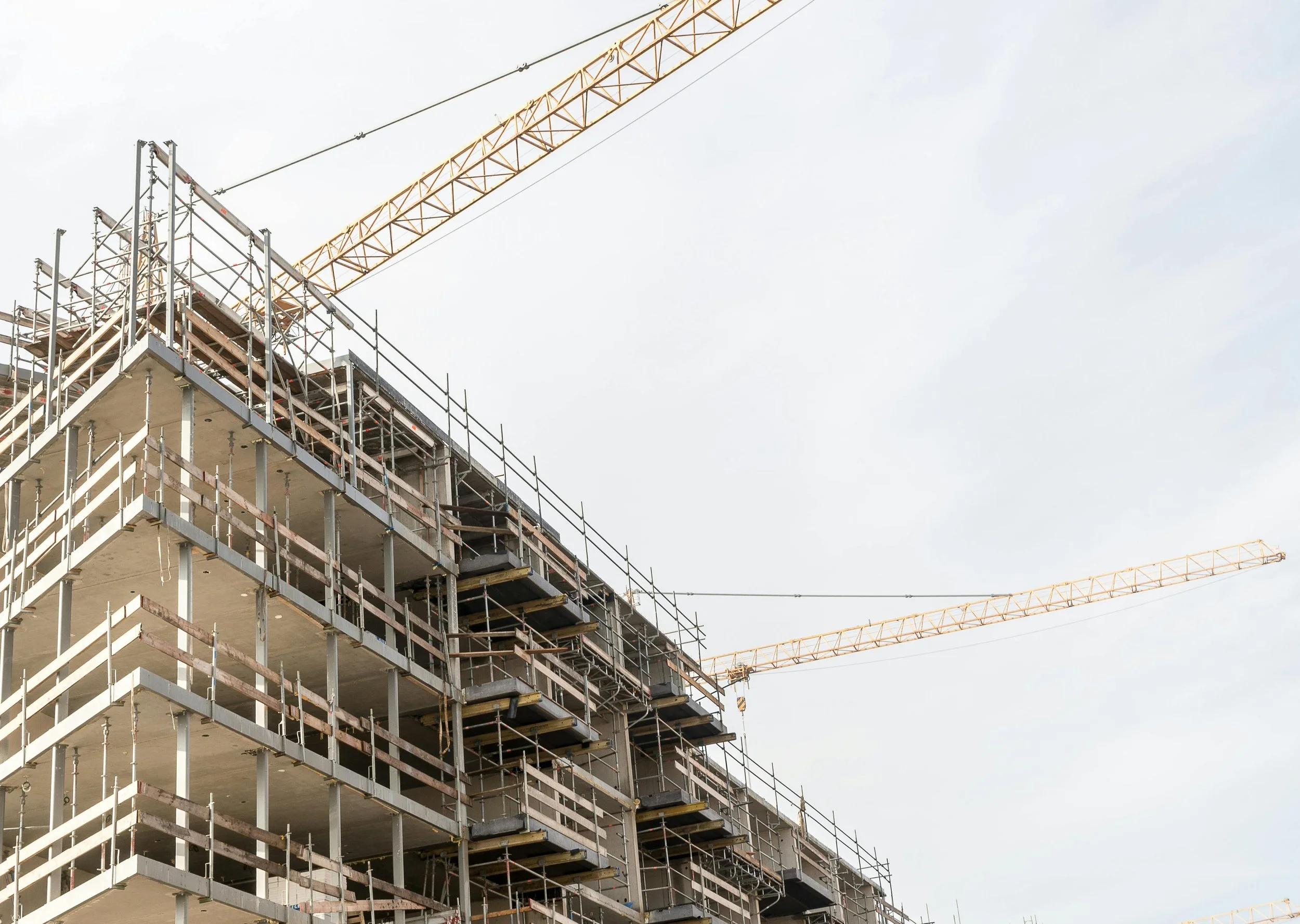 A construction site featuring a multi-story building under construction with scaffolding and two large yellow cranes above, set against a cloudy sky.