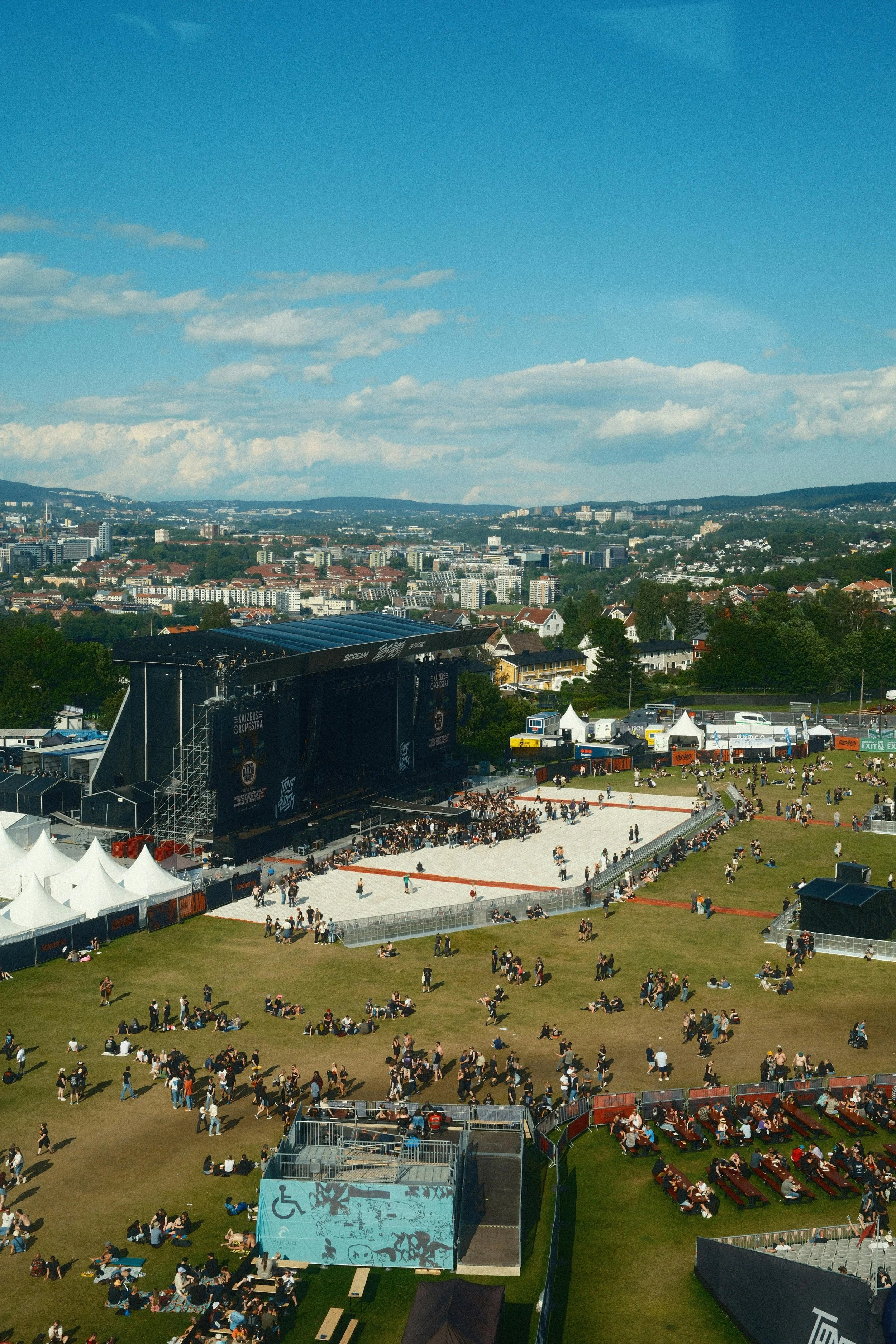An outdoor concert or festival with a stage, screened with black banners and tents, and a grassy area filled with people sitting and walking. In the background, there is a cityscape with buildings and hills under a partly cloudy sky.