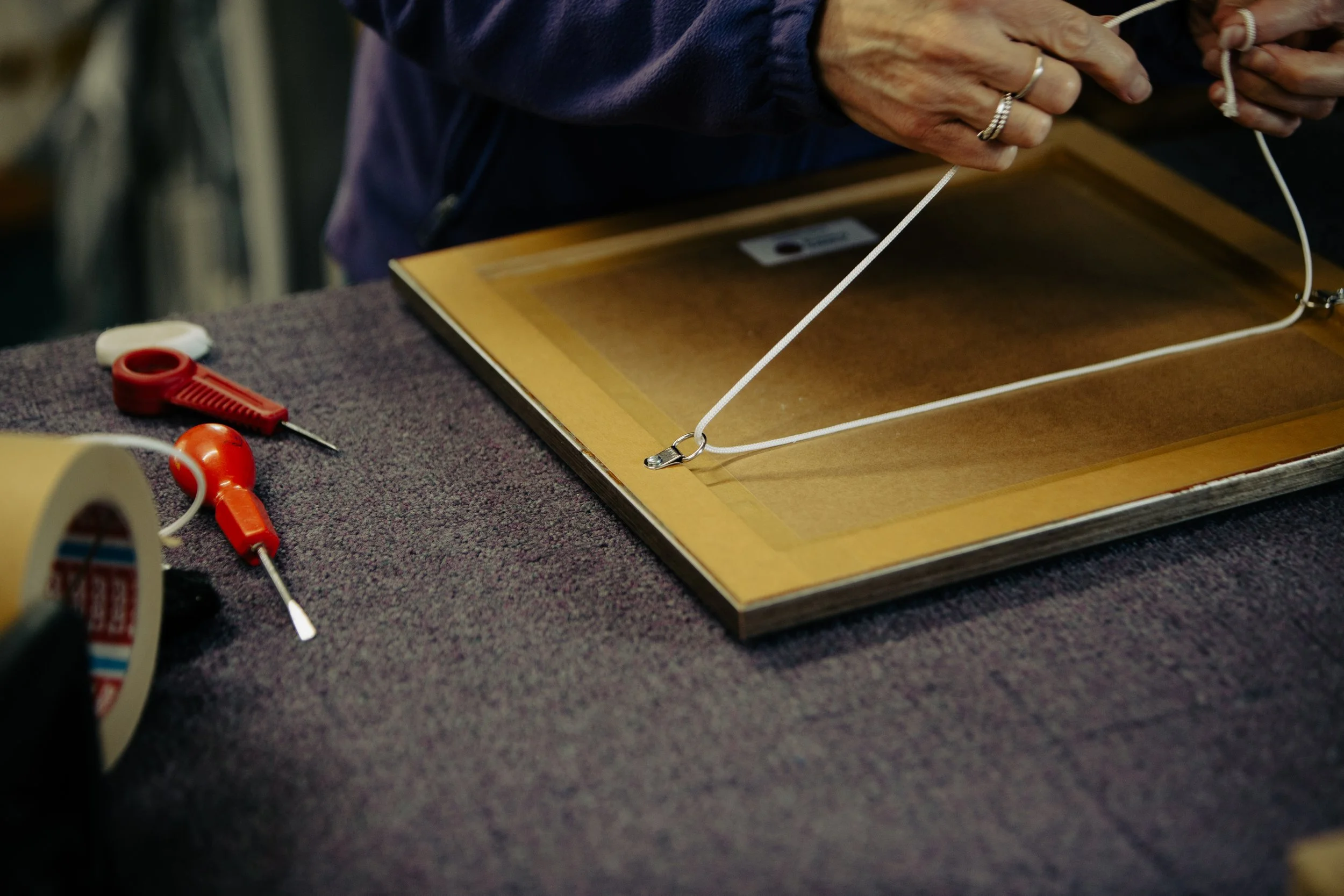 A person working on a framed project, using a nylon string attached to a metal ring on the frame, with tools like scissors and a screwdriver on the table.