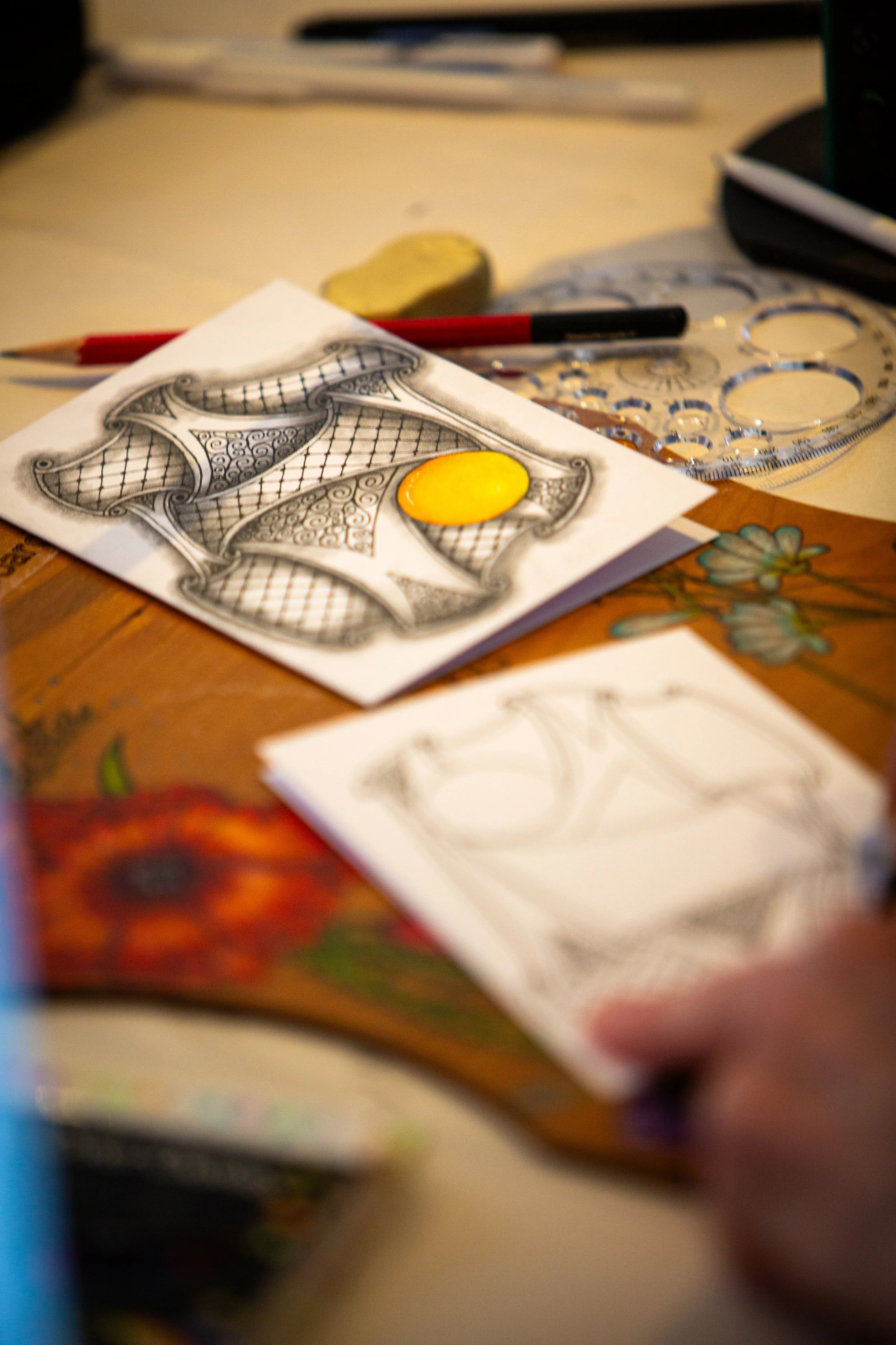 Close-up of a desk with a drawing of a decorative chair with an empty yellow circle on it, surrounded by a red pen, a black pen, a clear ruler, and a yellow eraser.
