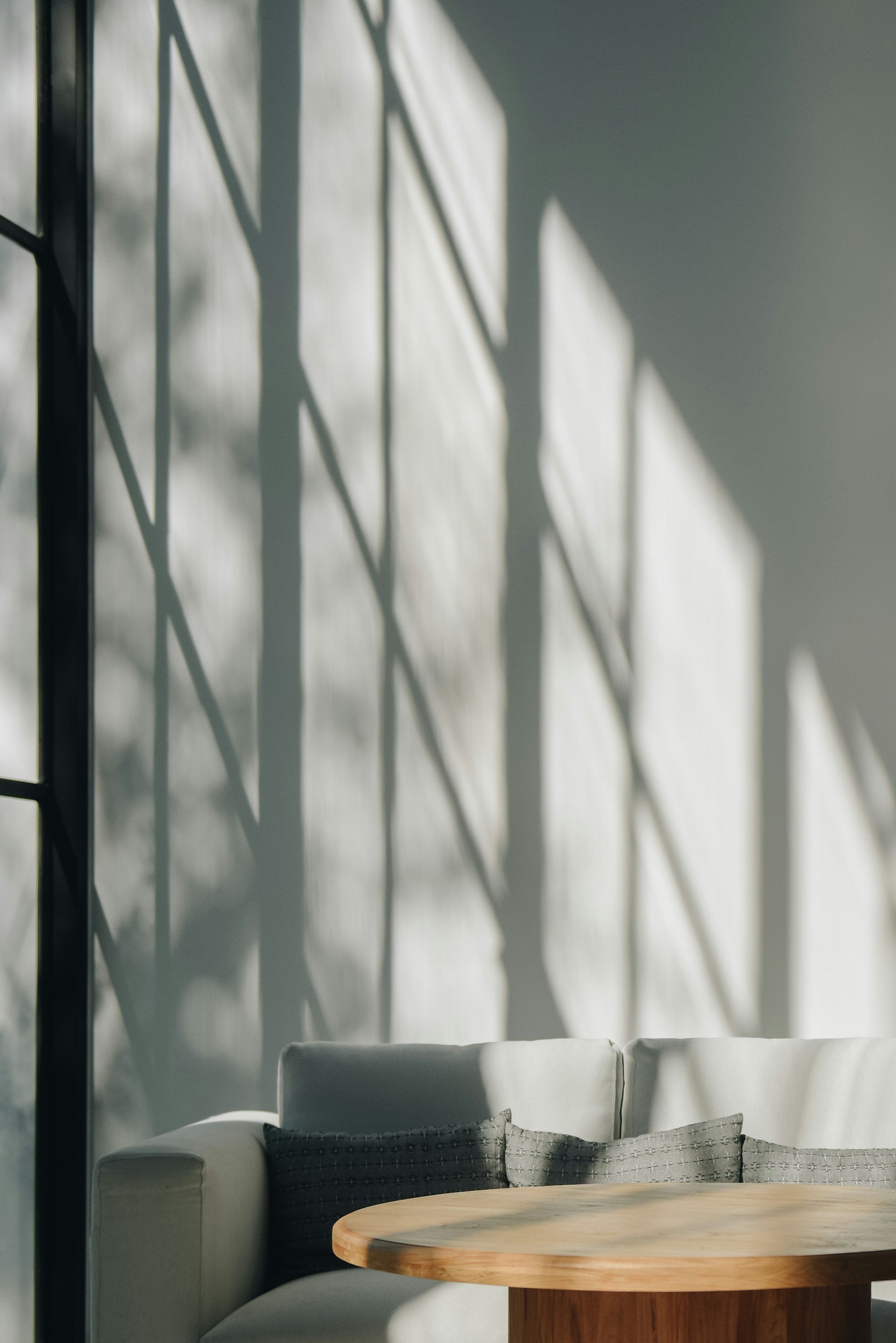 A cozy indoor space with a round wooden table, a light-colored sofa with a dark cushion, and large windows casting natural light and shadows on the wall.