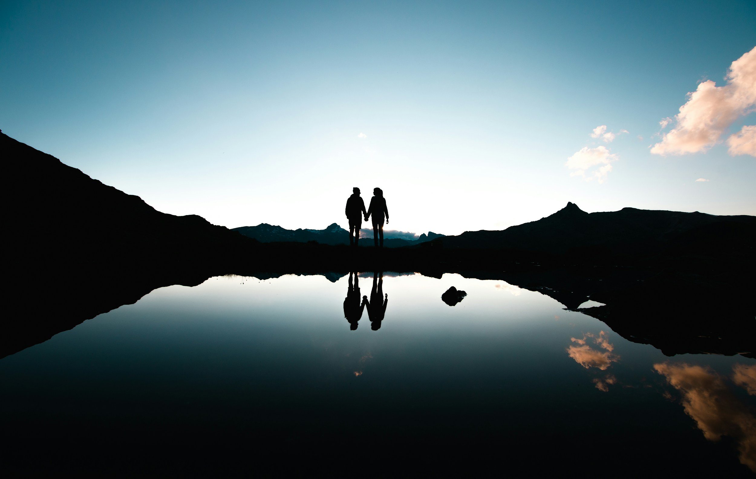 Two people holding hands walking along a mountain landscape during sunset, with their reflection visible in a calm body of water in the foreground.