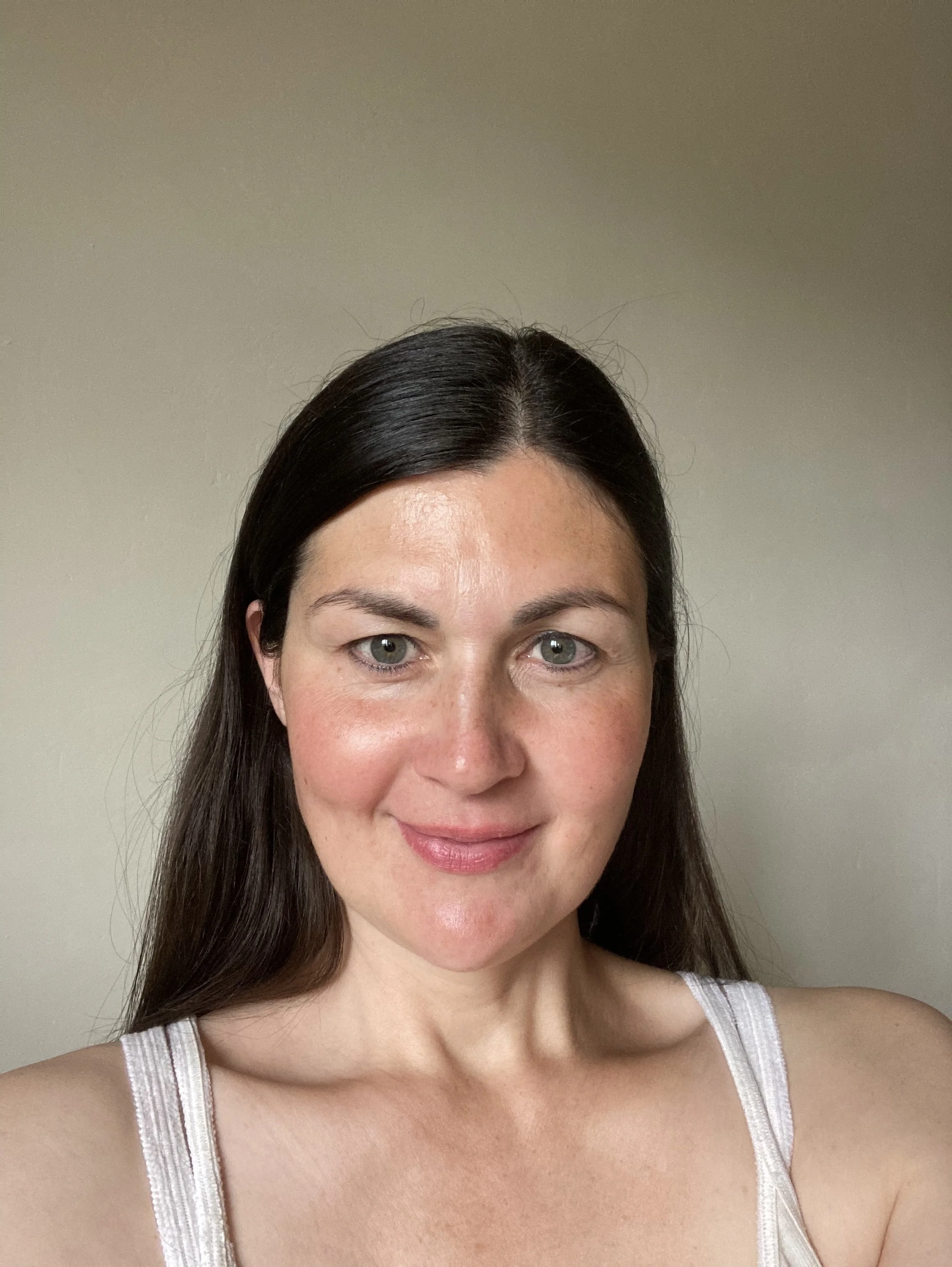 A woman with dark brown hair and blue eyes wearing a white tank top smiling at the camera against a plain beige wall.