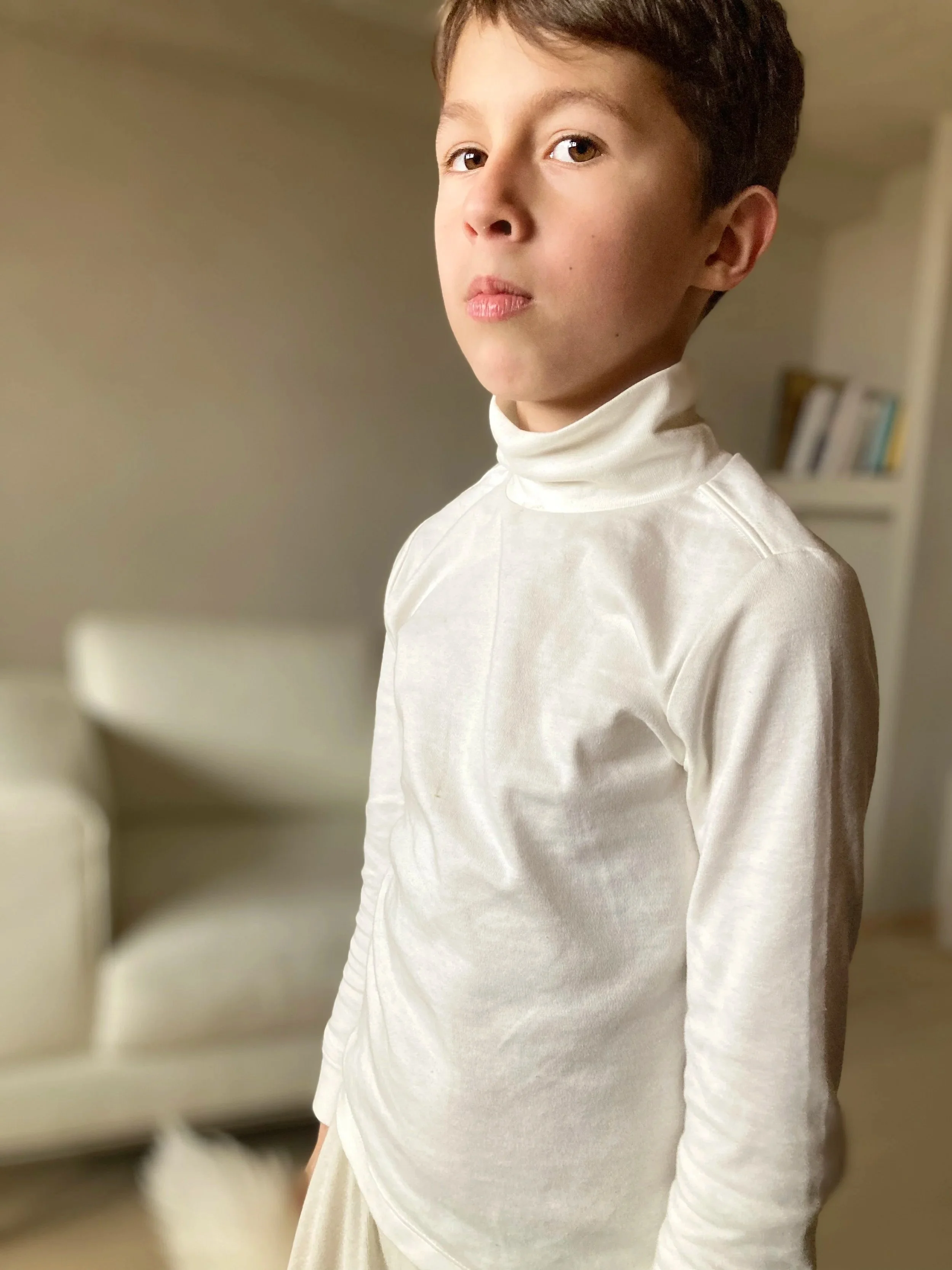 A young boy with short brown hair and light skin looking at the camera, wearing a white turtleneck shirt, standing in a room with a blurred background including a white bookshelf and some books.