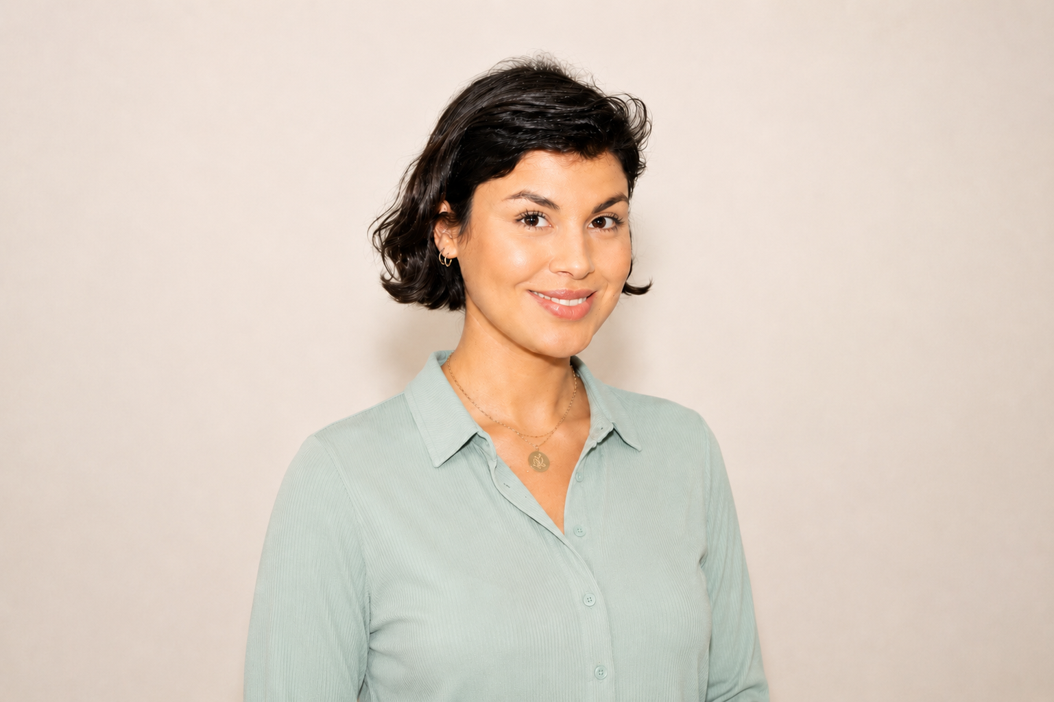 Junge Frau mit dunklen, lockigen Haaren trägt eine hellblaugrüne Bluse und eine goldene Halskette, lächelnd gegen einen hellen Hintergrund.