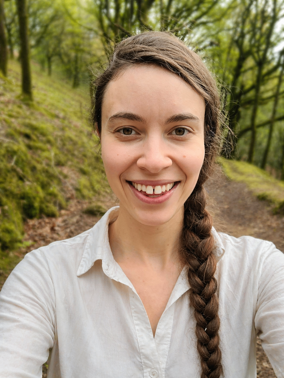 A woman with long brown hair in a braid, smiling, taking a selfie in a green forested area.