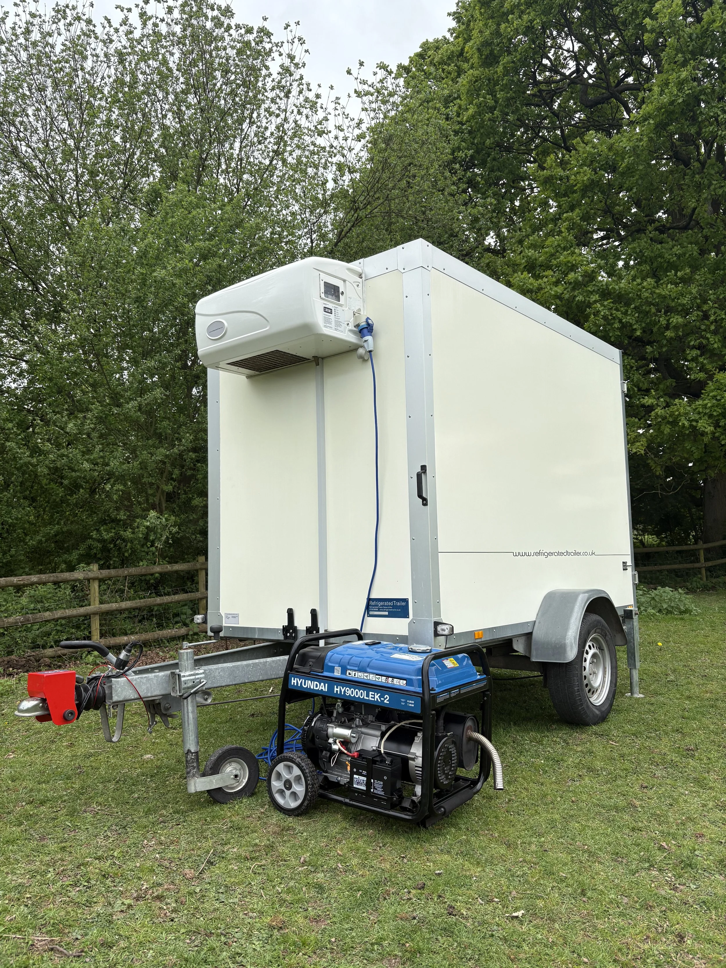 A mobile refrigerated trailer connected to a generator and cooling unit, set up outdoors on grass with trees in the background.