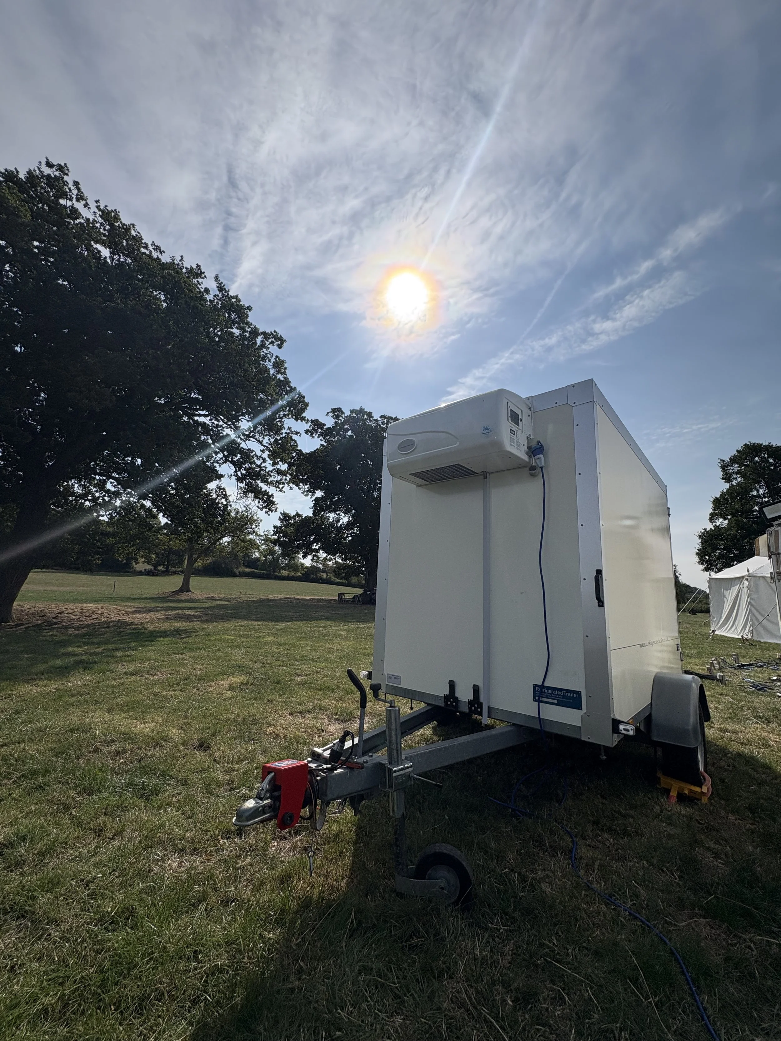 A white refrigerated trailer with an air conditioning unit mounted on top, parked outdoors on grass, under a partly cloudy sky with the sun shining brightly.