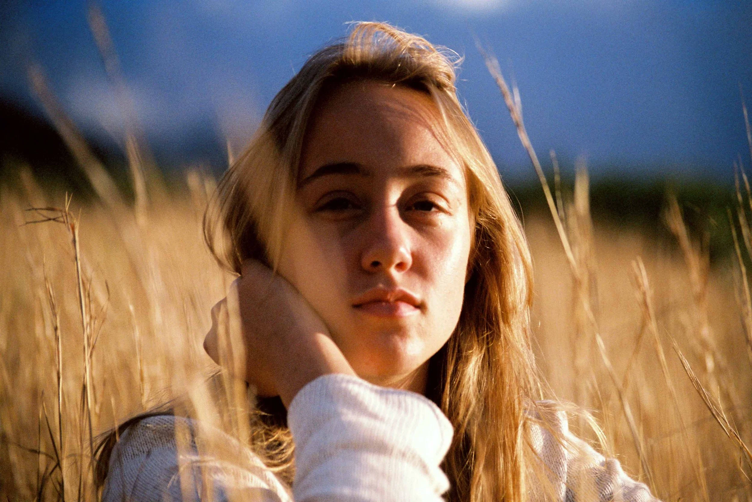 A young woman with long, wavy hair standing in a field of tall, golden grass under a clear blue sky, looking into the distance with a relaxed expression.