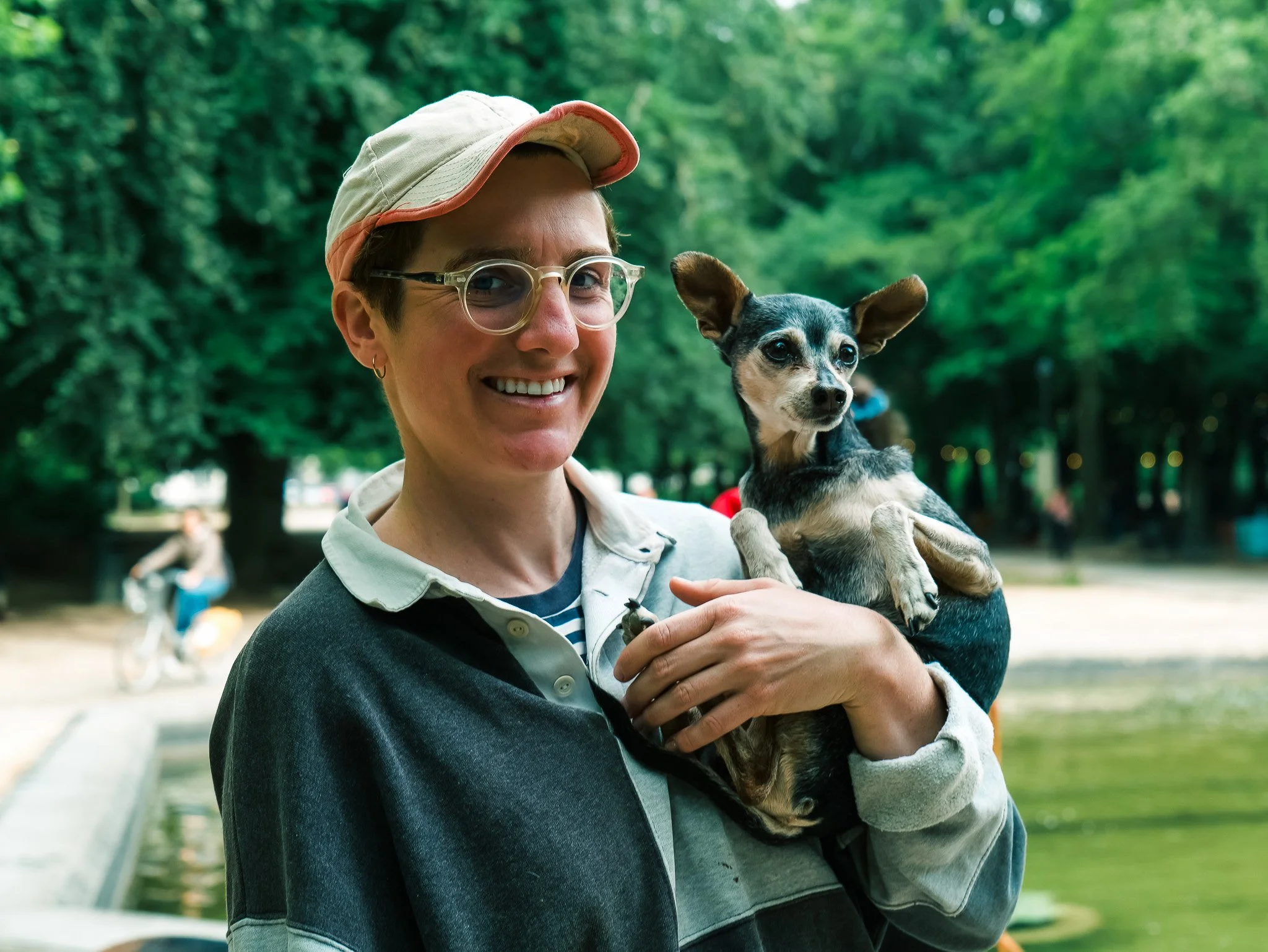 A woman wearing glasses, a beige hat with a pink trim, and a dark sweater holds a small dog with a black and tan coat outdoors in a park with green trees.