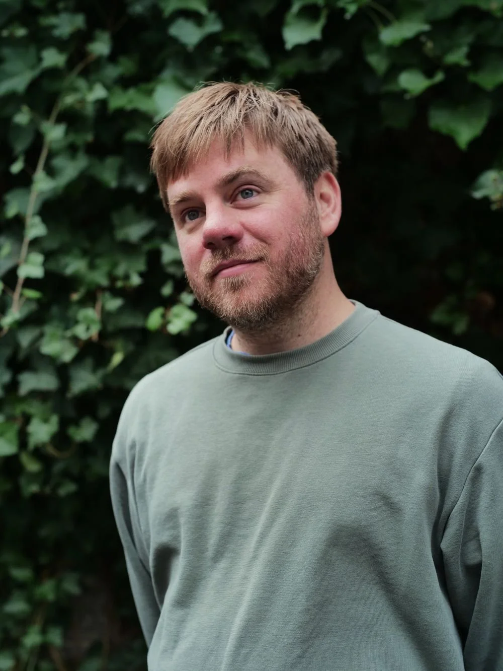 A man with light brown hair and a beard, wearing a green sweatshirt, stands outdoors in front of green leafy bushes.