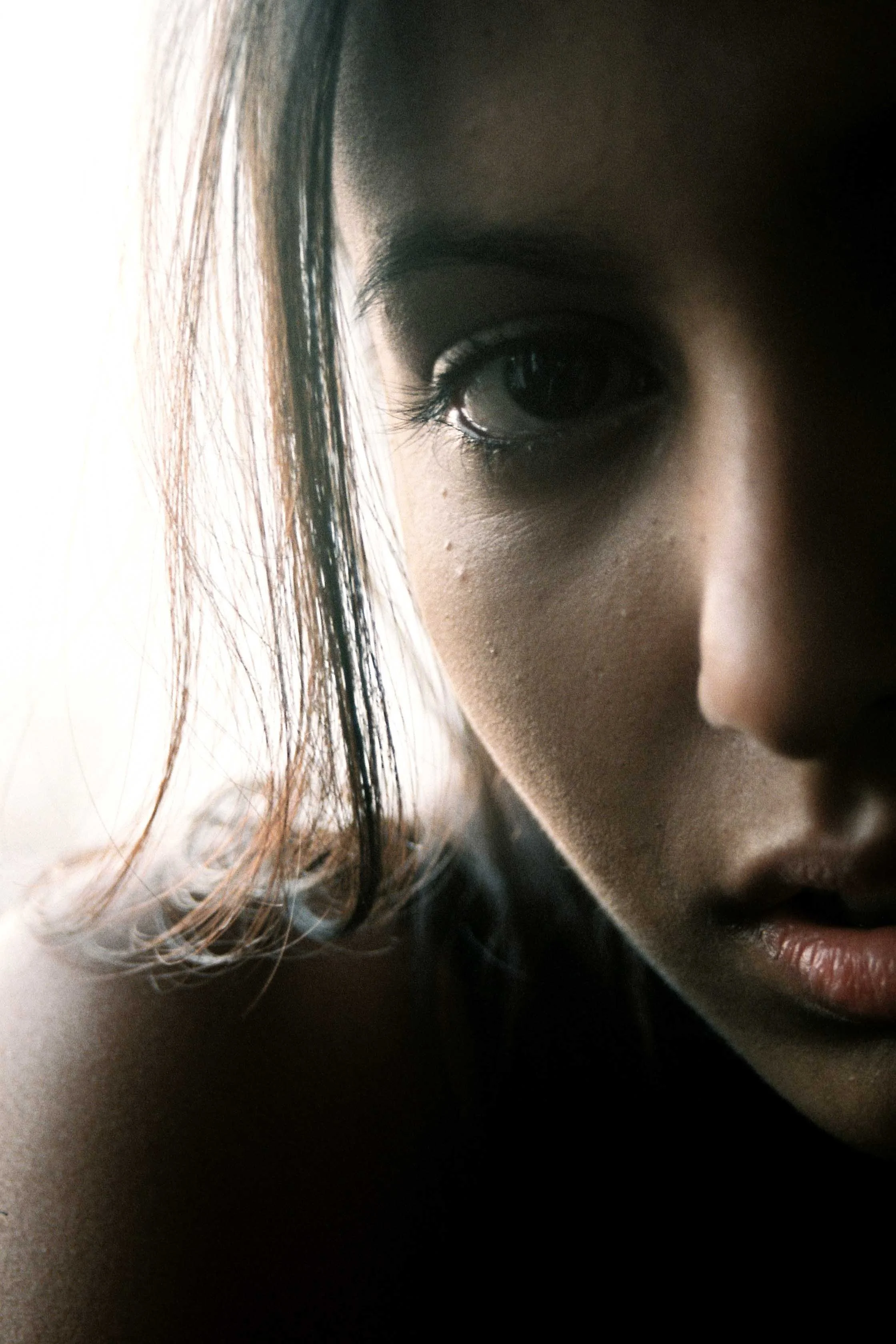 Close-up of a person's face with dark hair and eye, partially obscured by shadows, showing a pensive expression.